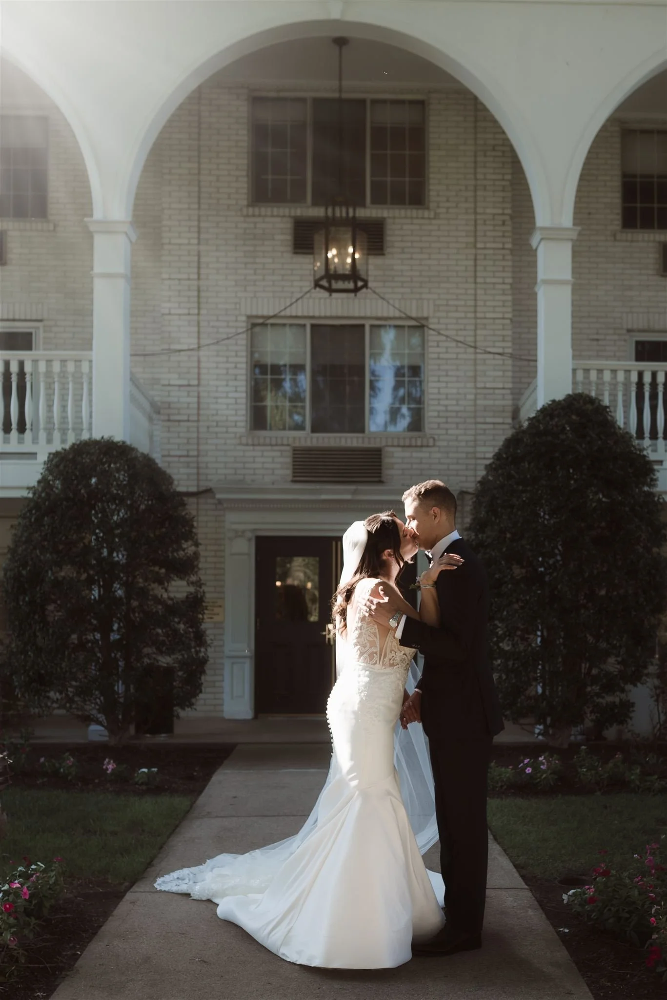 Bride and groom sharing a kiss outside a building with arched entryway and brick facade.