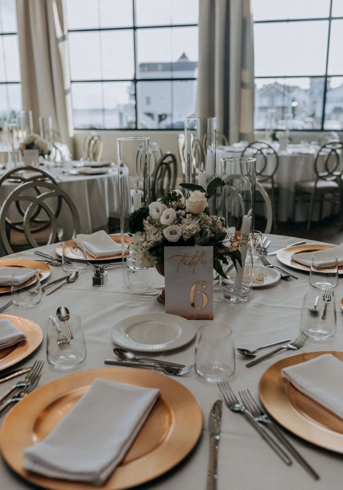 Round banquet table set for a wedding reception, with white tablecloth, gold chargers, white napkins, wine and water glasses, silverware, floral centerpiece, candle holders, and a table number card displaying the number 6. Large windows in the backgr