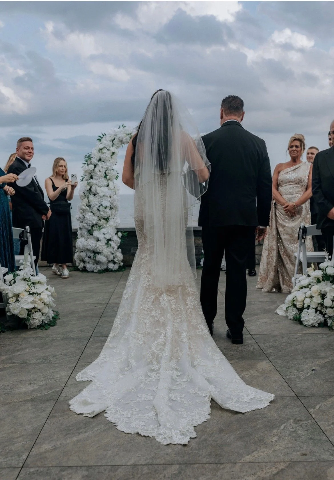 A bride and groom standing at the altar during outdoor wedding ceremony with cloudy sky, surrounded by friends and family, with floral decorations and an ocean view in the background.