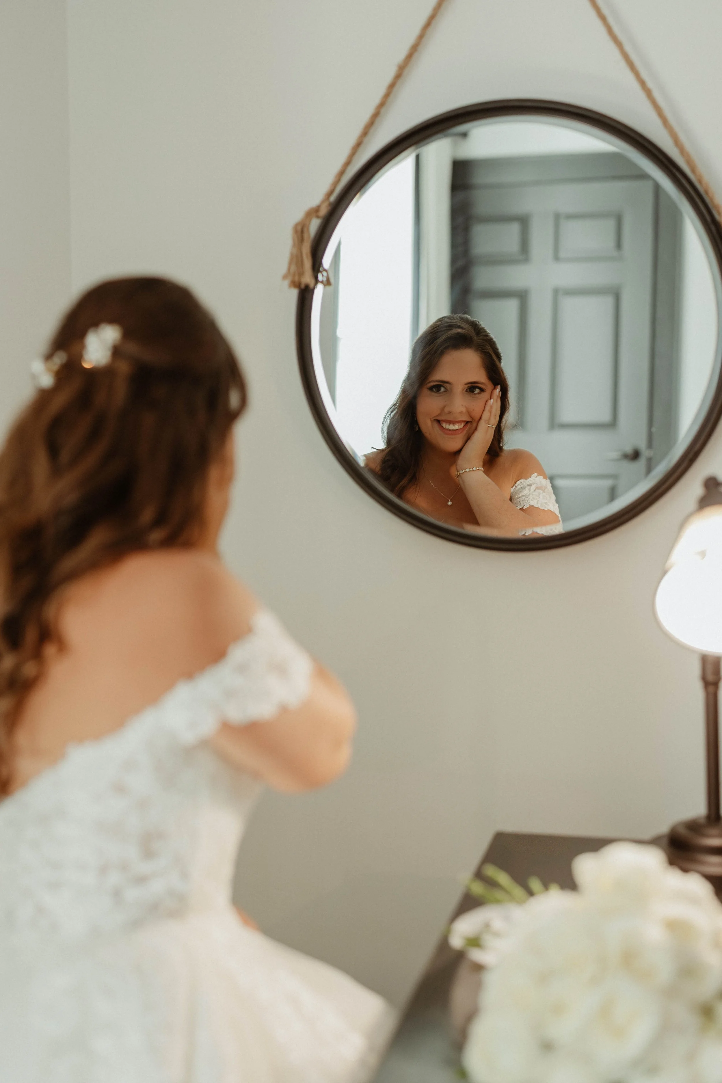 Bride smiling at herself in a round wall mirror while touching her face, with a white wedding dress and floral hair accessory, on a dressing table next to a bouquet of white roses.