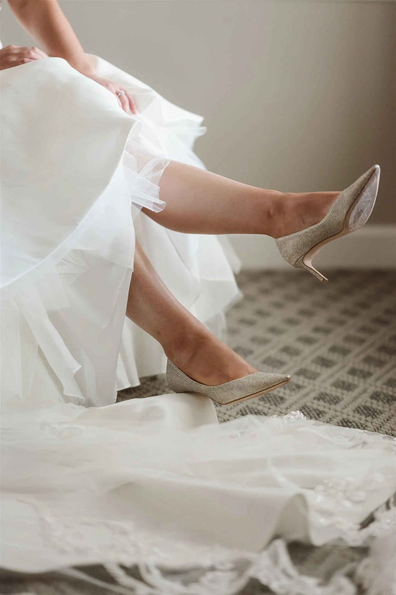 A bride in a white wedding dress adjusting her champagne-colored high heels, with the dress slightly lifted to reveal one shoe and part of her leg on a patterned carpet.