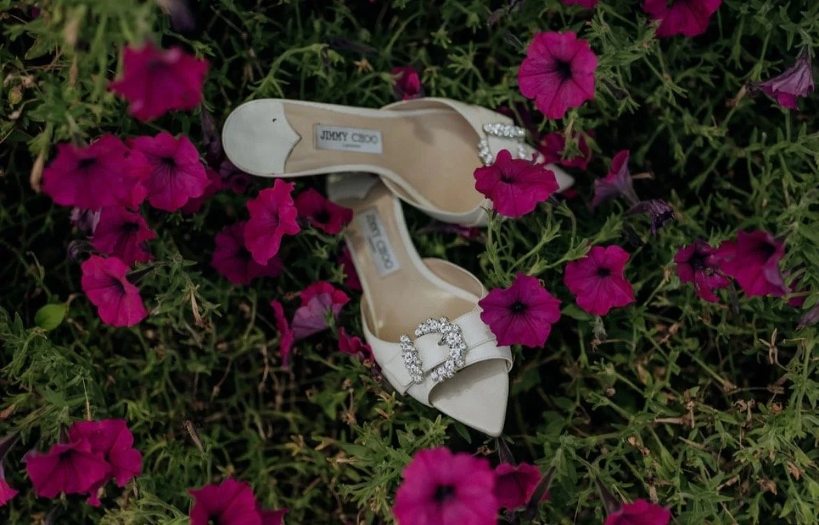 White Jimmy Choo high-heeled shoes with rhinestone embellishments on the straps, lying among pink and purple petunias in a garden.