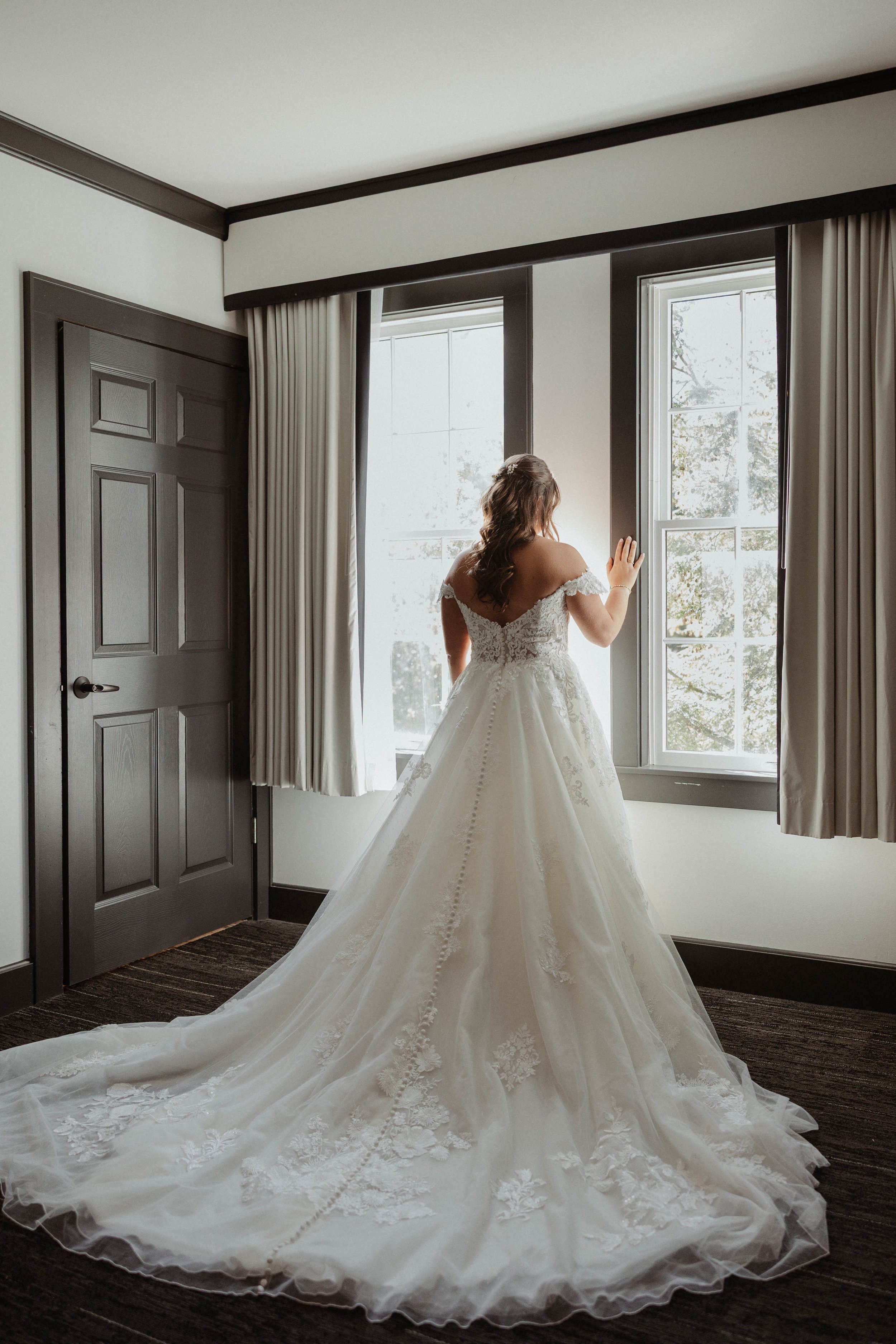 A bride in a white wedding gown standing by a window, gazing outside, in a room with dark wood trim and a closed door.