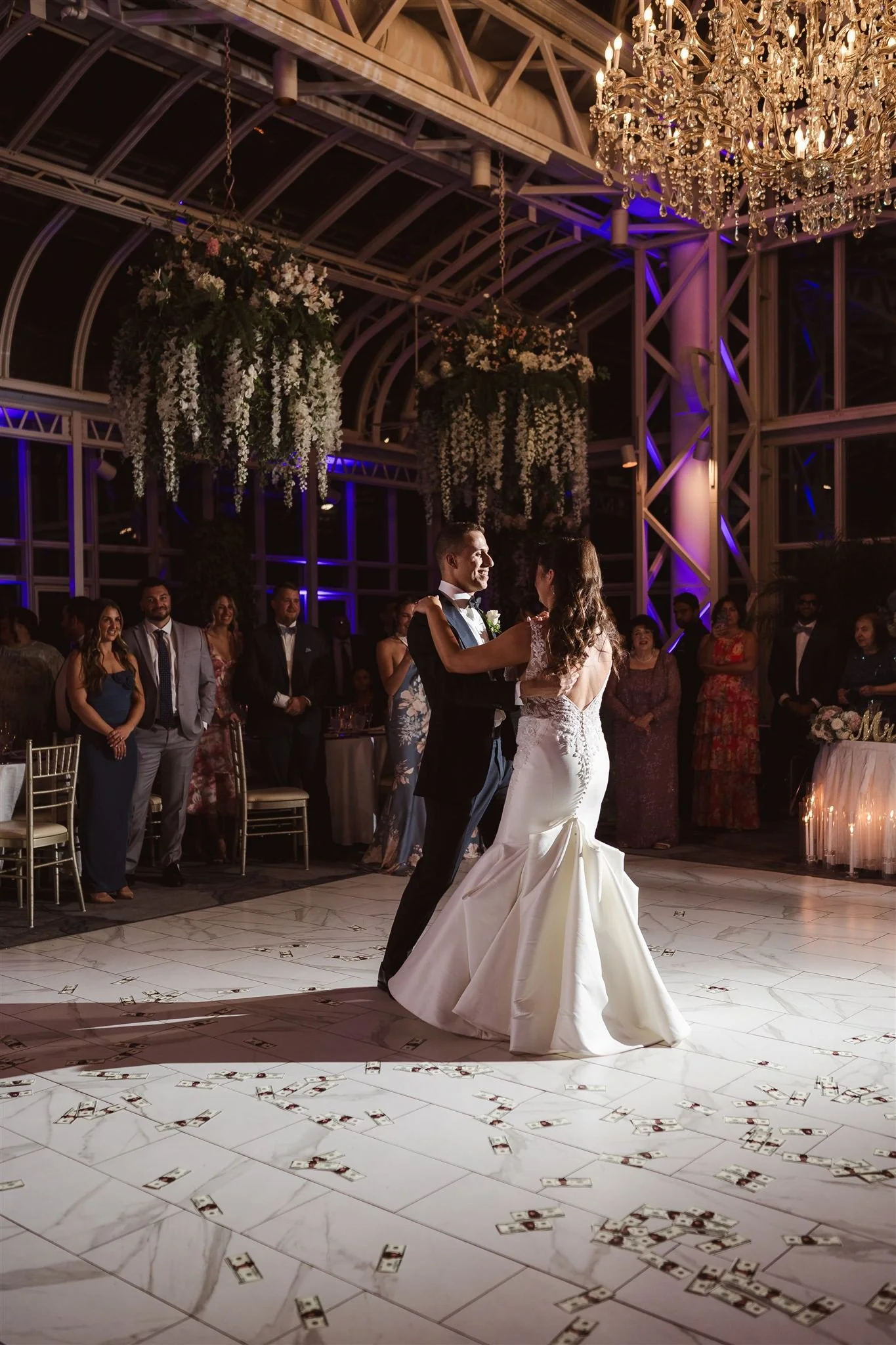 A bride and groom dancing in the center of a wedding reception hall with guests watching, money scattered on the dance floor, large hanging floral arrangements, and a crystal chandelier overhead.