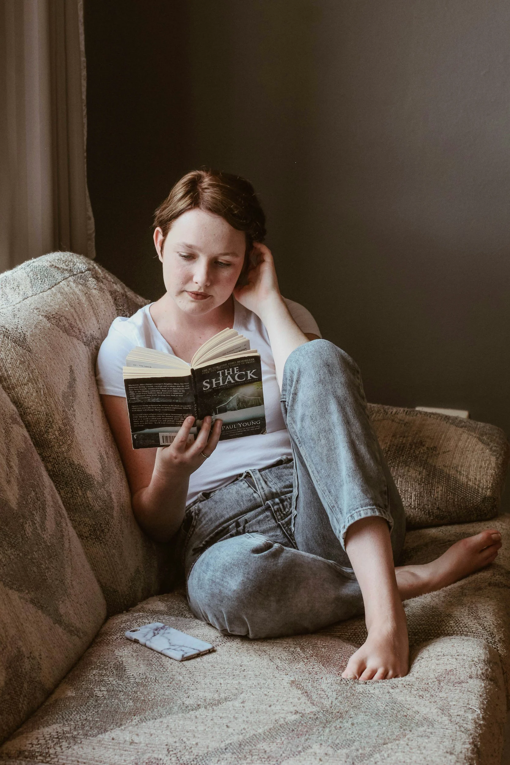 A young woman with short brown hair reading a book titled 'The Shack' while sitting on a beige couch, with a phone on the couch beside her.