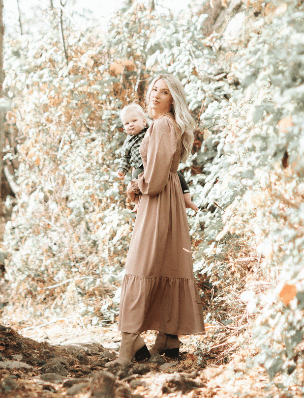 A woman with long blonde hair holds a young child in a wooded area with autumn leaves. The woman wears a tan dress and boots, and the child wears a checkered shirt. They are standing on a forest trail surrounded by trees with orange and green foliage.