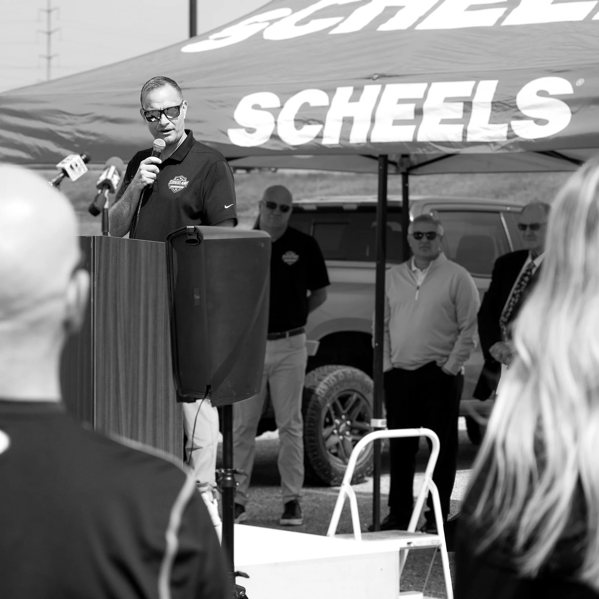 A man speaks into a microphone at an outdoor event under a tent with the brand 'SHEL' visible. Several people stand behind him, with a vehicle visible in the background.
