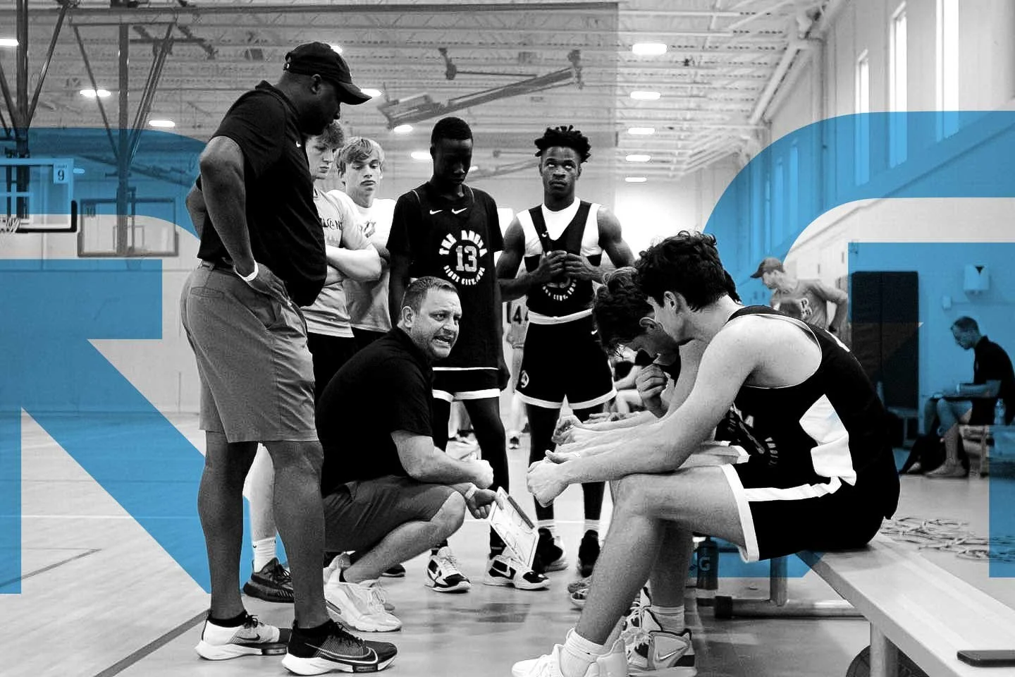 A coach giving instructions to a youth basketball team during a timeout on the sidelines of a basketball court, with players listening intently, some seated and some standing.