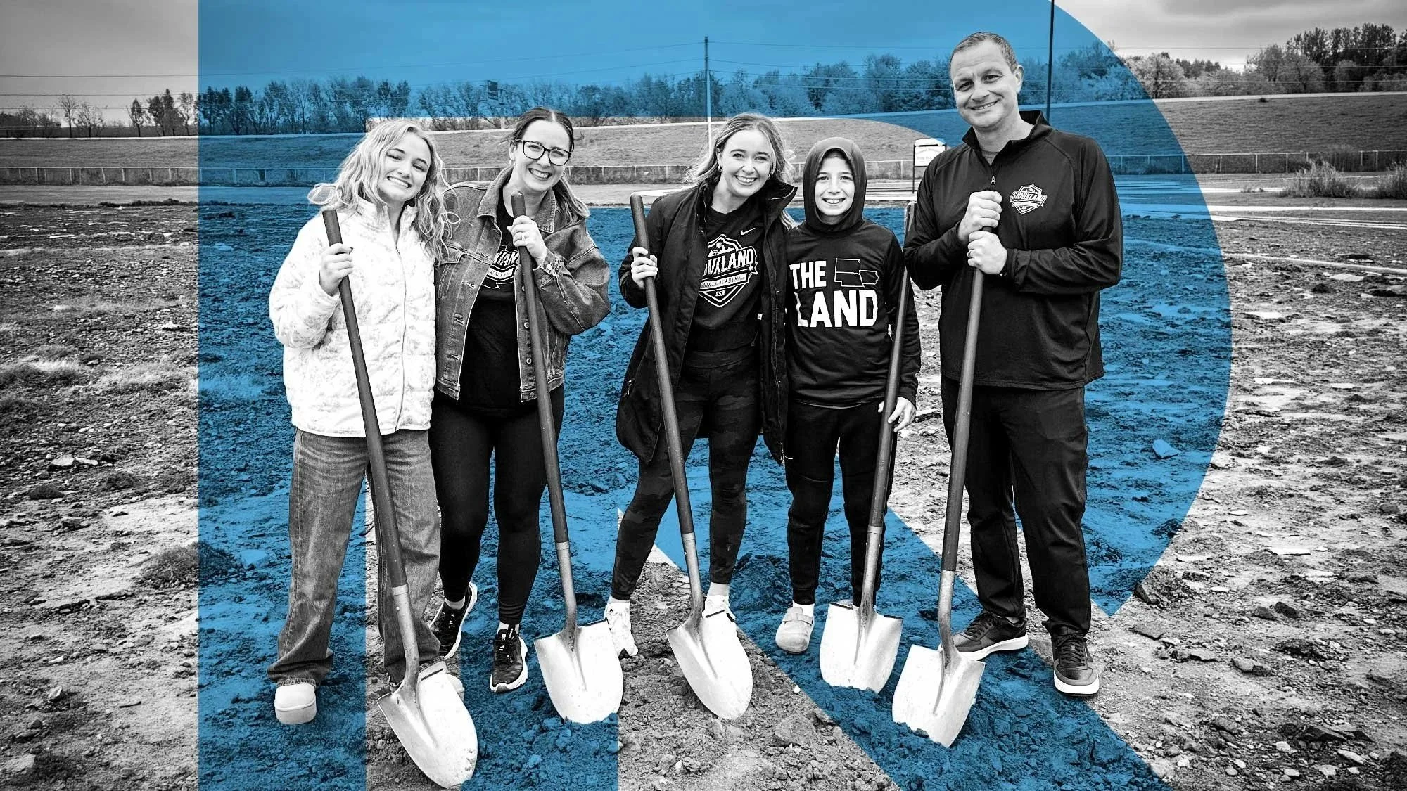 Group of five people standing on a dirt field holding shovels, smiling, with trees and a fence in the background. Image is in black and white with blue highlights.