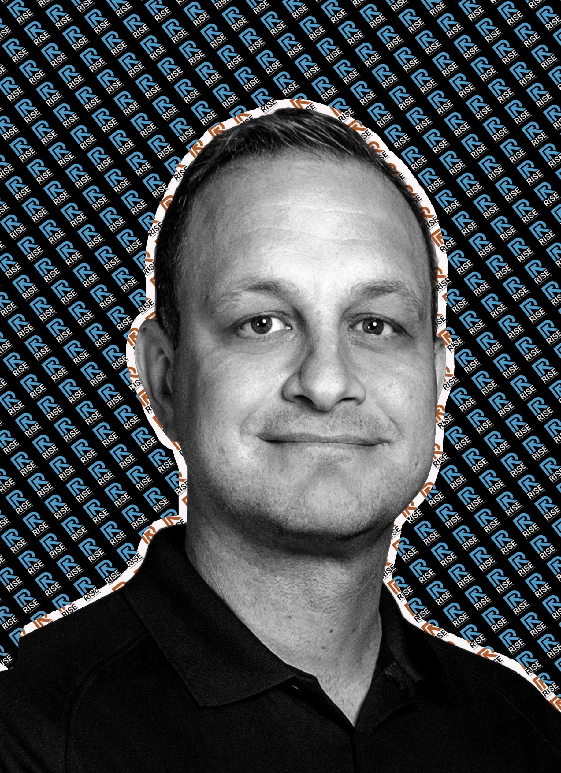 Black-and-white portrait of a man with short hair, smiling, with a background patterned with logos saying 'RISE' in blue and white.