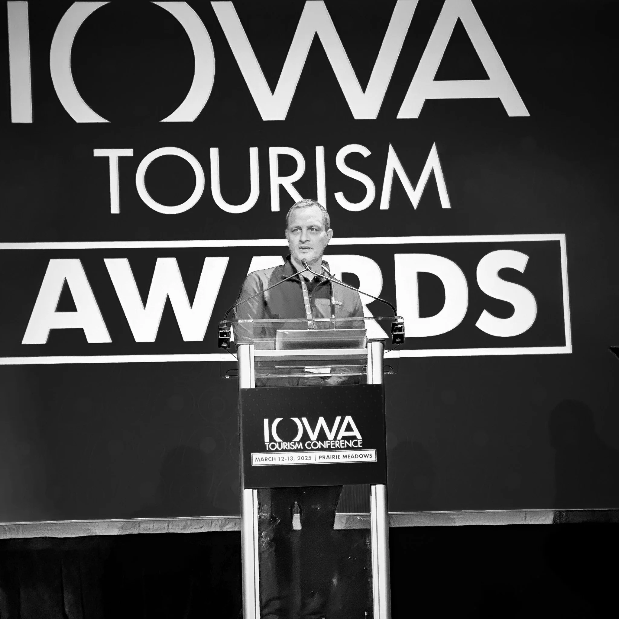 A man giving a speech at a podium during the Iowa Tourism Conference, March 12-13, 2025, at Prairie Meadows, with a large backdrop displaying the event's name.