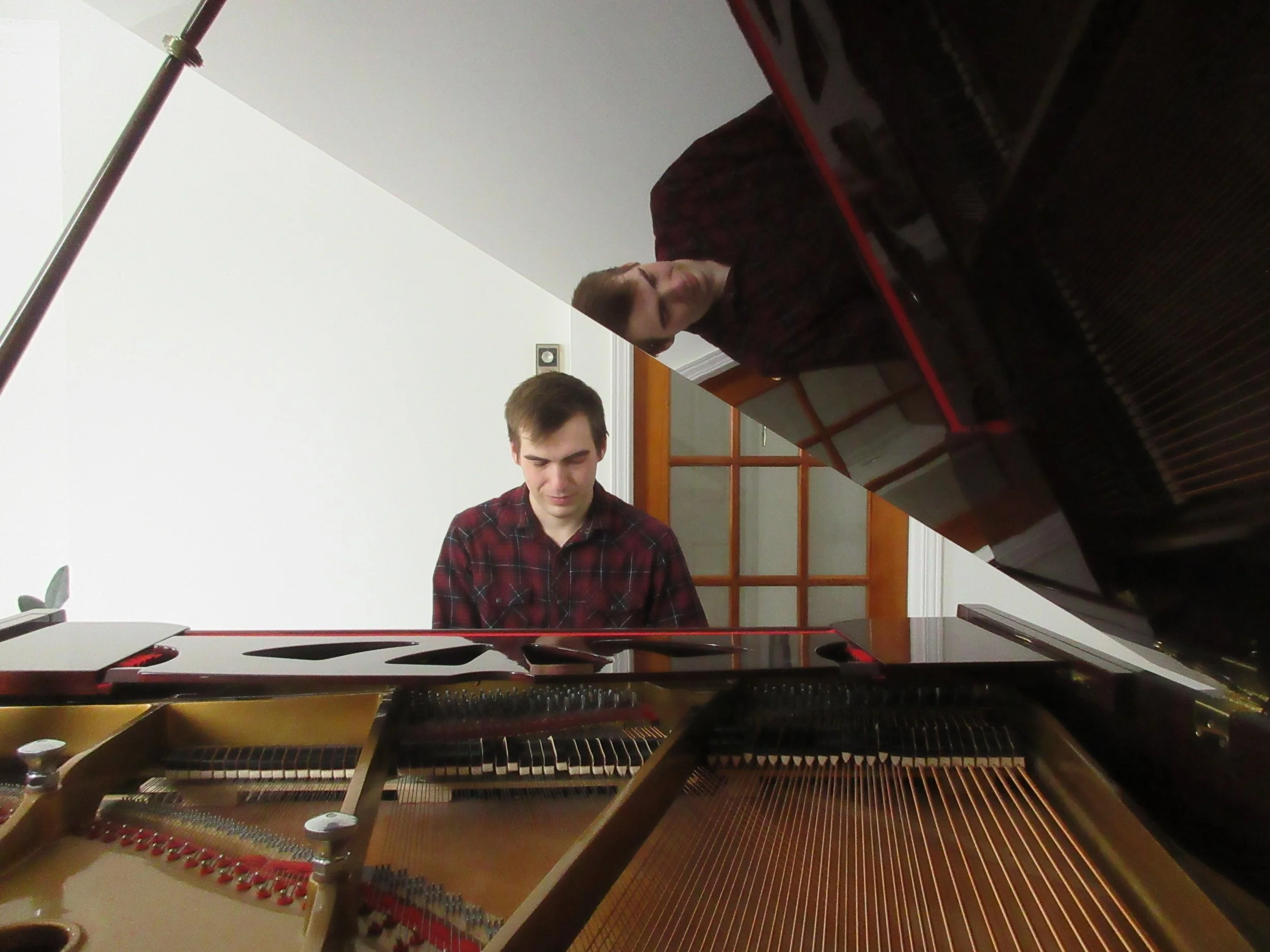 Young man playing a grand piano, viewed from inside the piano with the lid open, in a room with wooden door and white walls.