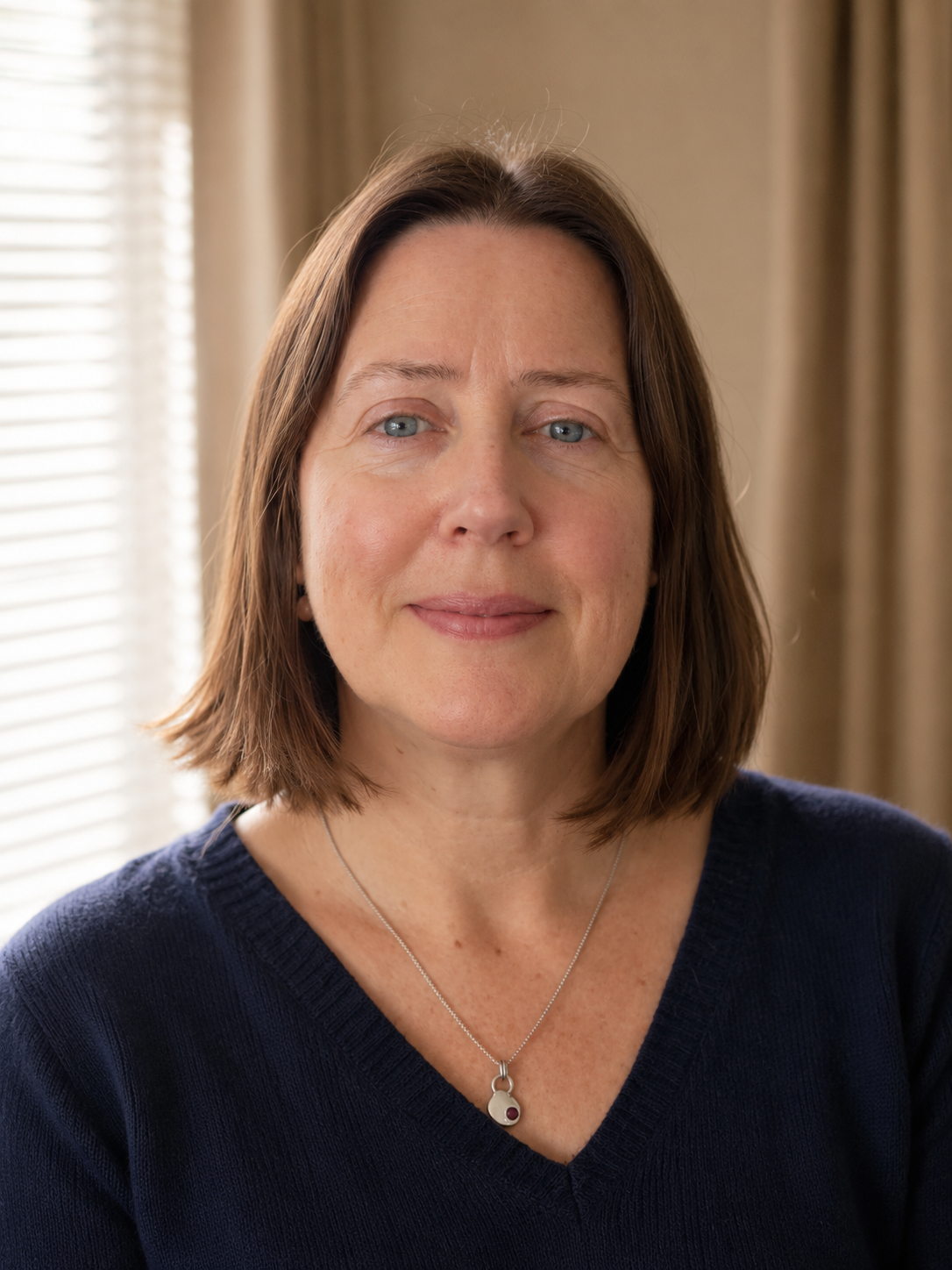 A middle-aged woman with shoulder-length brown hair and blue eyes wearing a navy sweater and a silver necklace with a small, round pendant, standing in front of beige curtains and blinds.