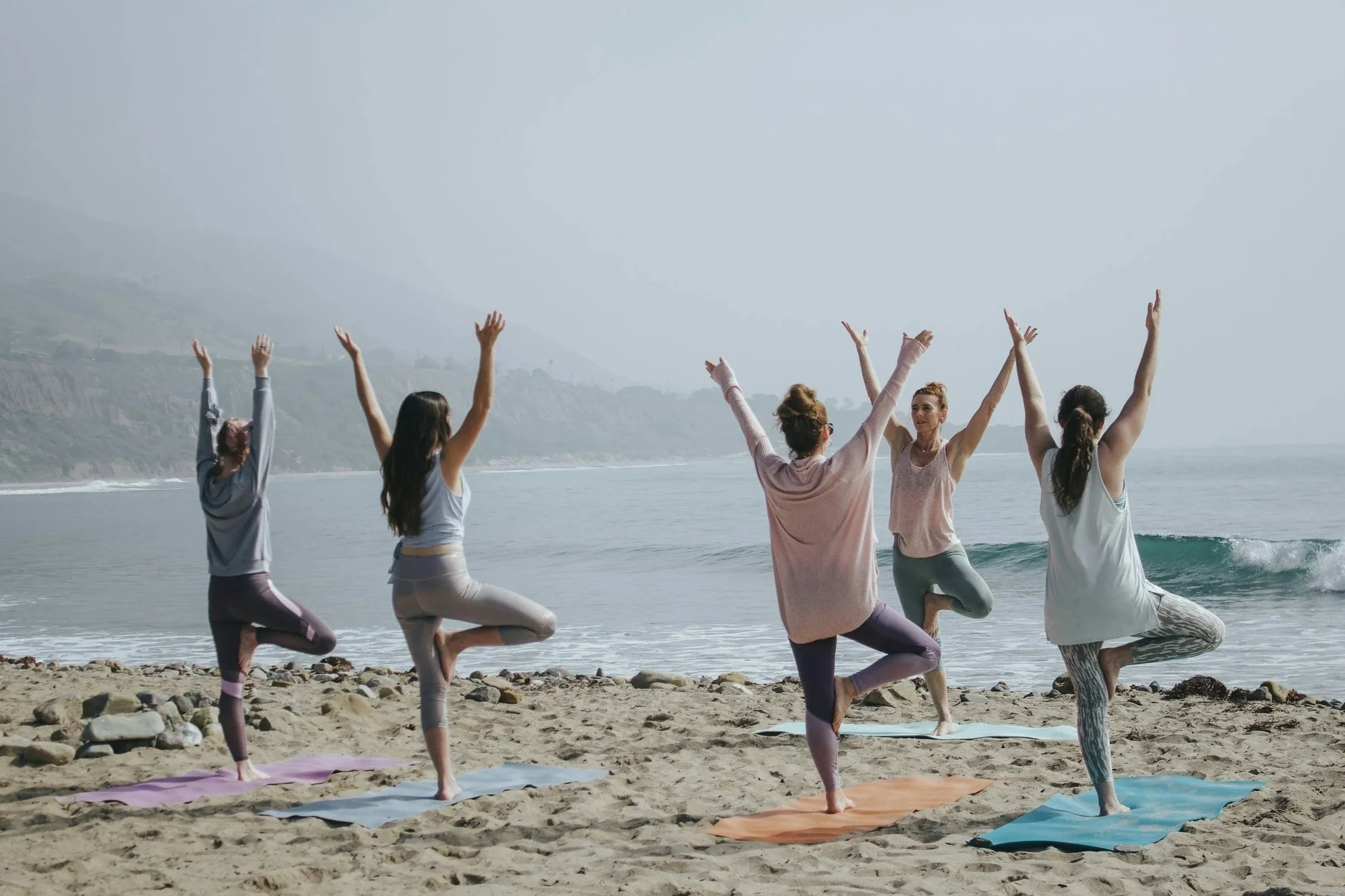 Five women practicing yoga on a beach with the ocean and hills in the background, performing tree pose with arms raised.