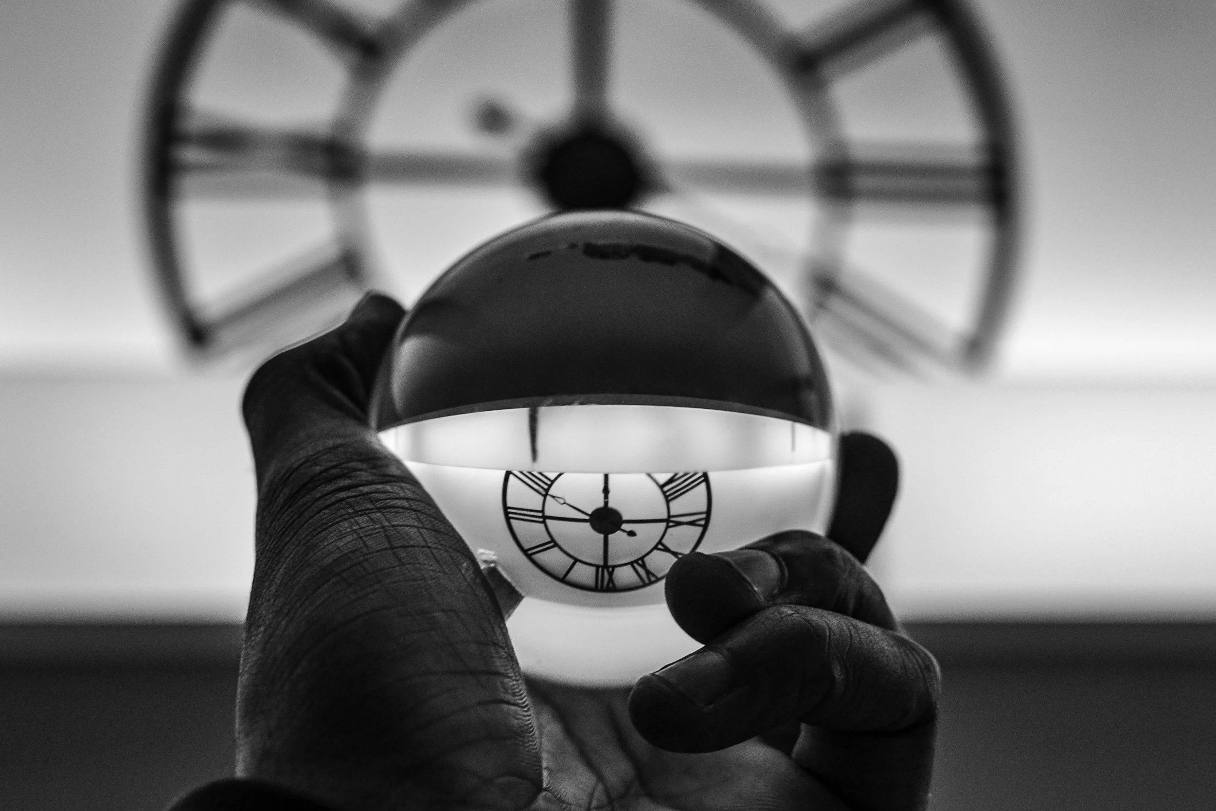 Black and white photo of a person's hand holding a glass sphere in front of a clock with Roman numerals, creating a reflection of the clock inside the sphere.