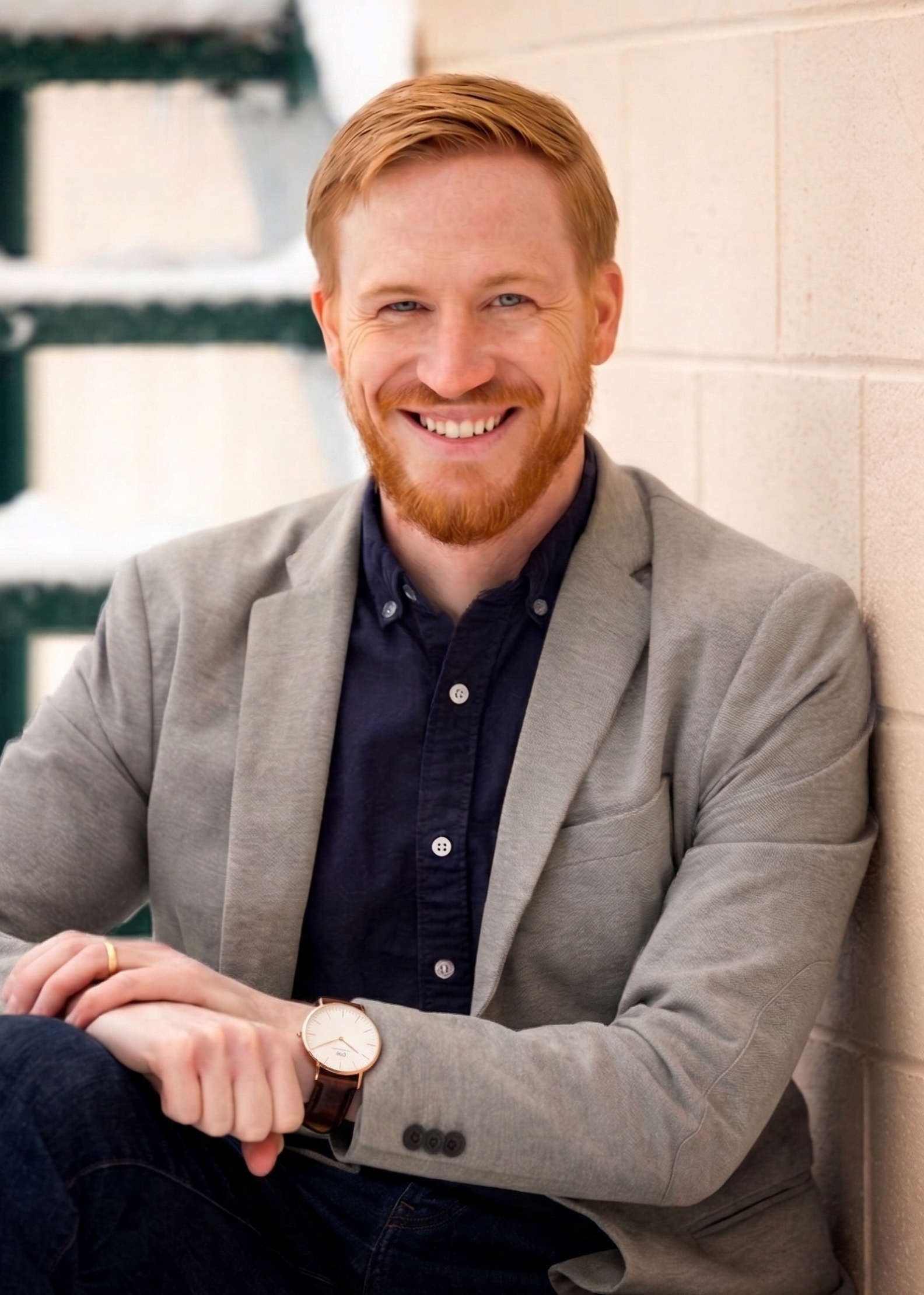 A smiling man with red hair and beard, wearing a gray blazer, navy shirt, and watch, sitting against a brick wall.