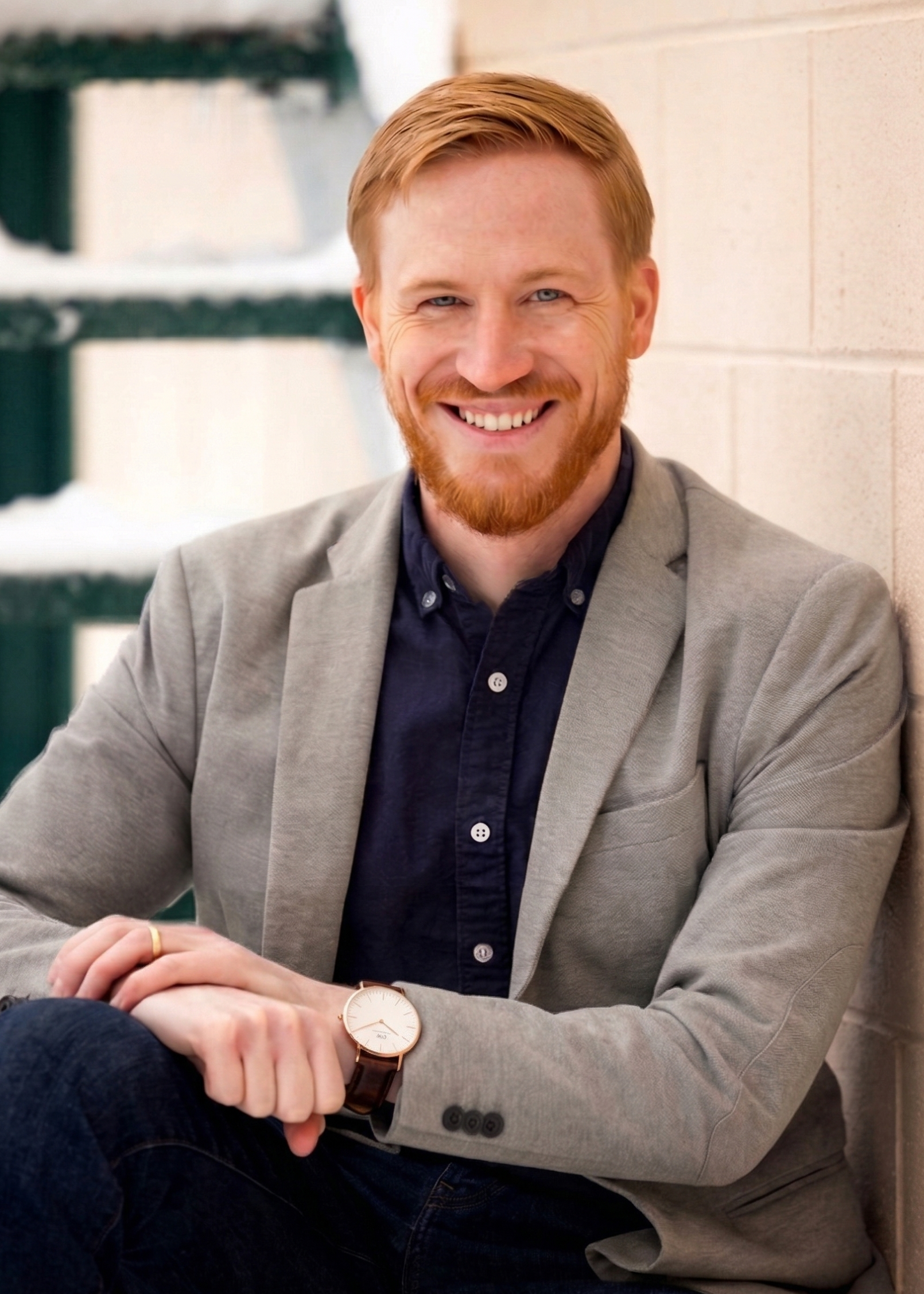 A smiling man with red hair and beard, wearing a gray blazer, dark shirt, and a watch, sitting against a brick wall with a staircase in the background.