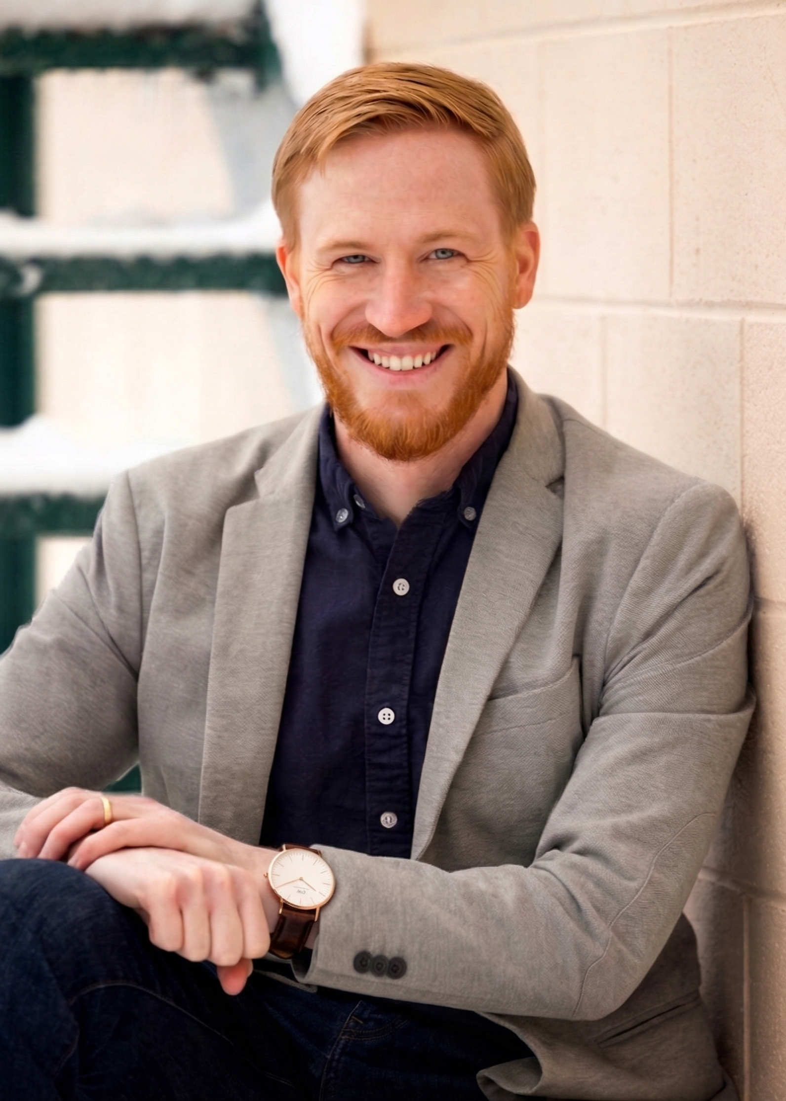 A smiling man with red hair and a beard, wearing a gray blazer and a dark button-up shirt, sitting against a brick wall with a staircase in the background.