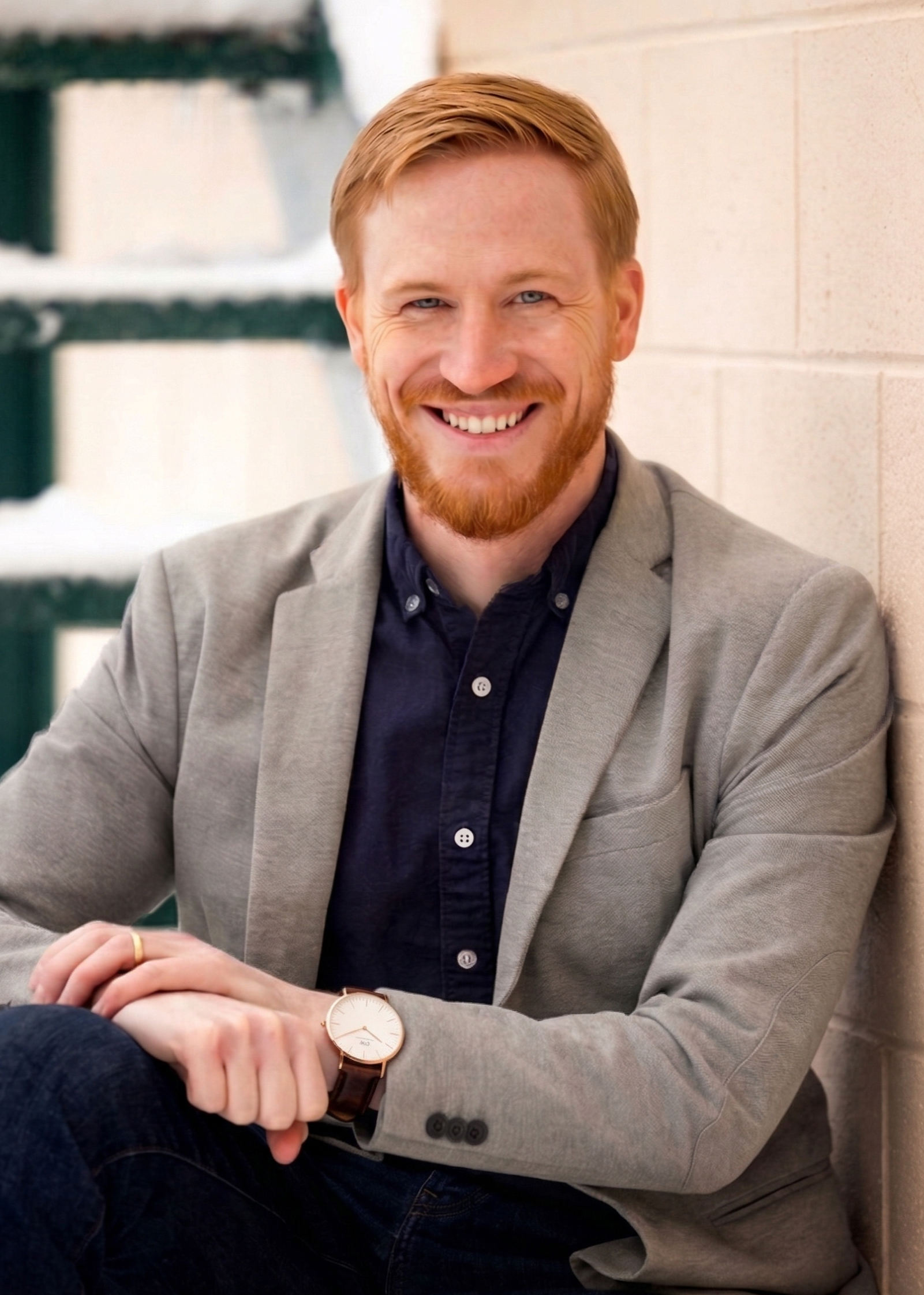 A smiling man with red hair and beard, wearing a gray blazer and dark shirt, sitting against a beige brick wall.
