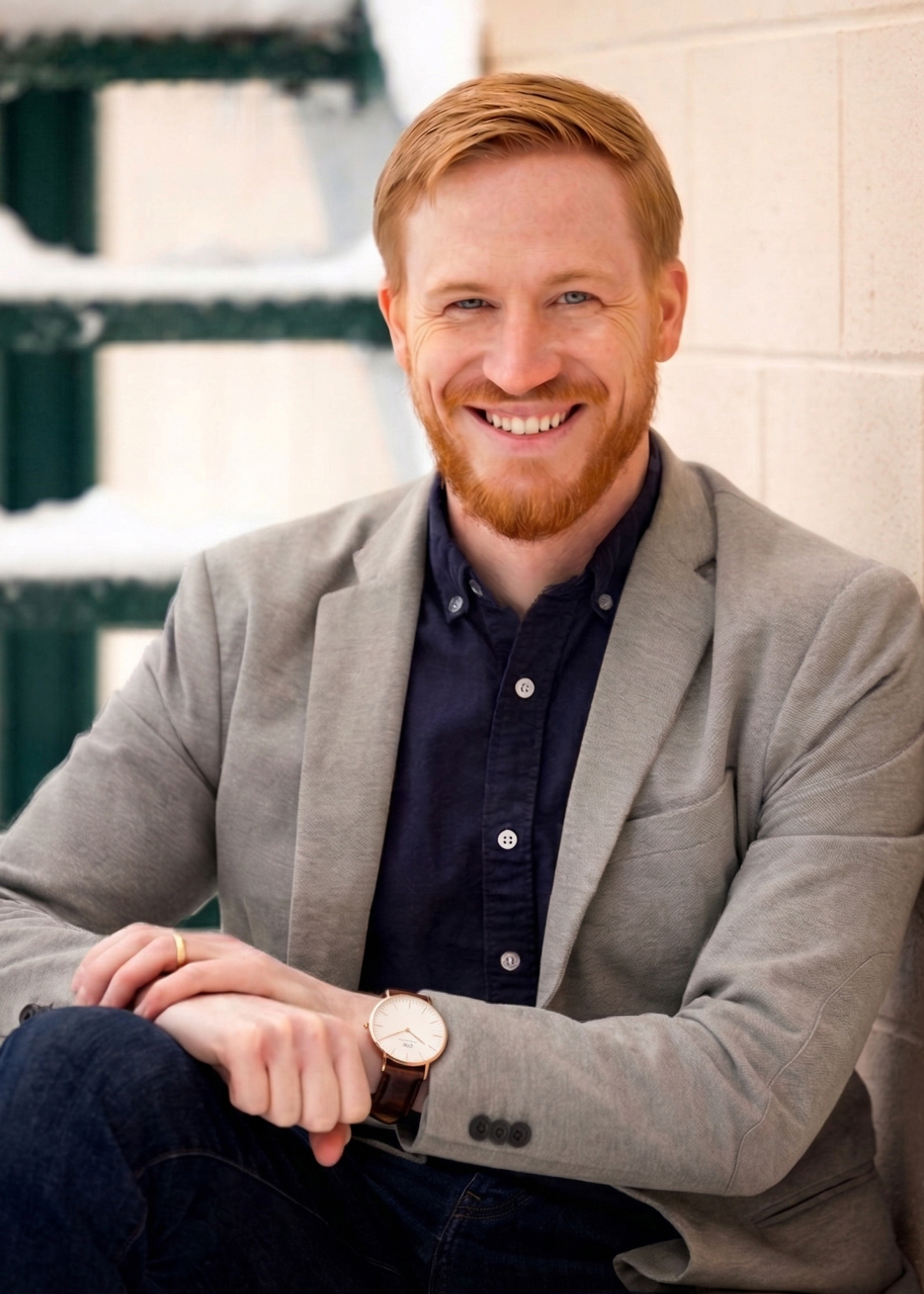 Smiling man with red hair, beard, wearing a gray blazer and dark shirt, sitting outdoors near a wall and stairs.