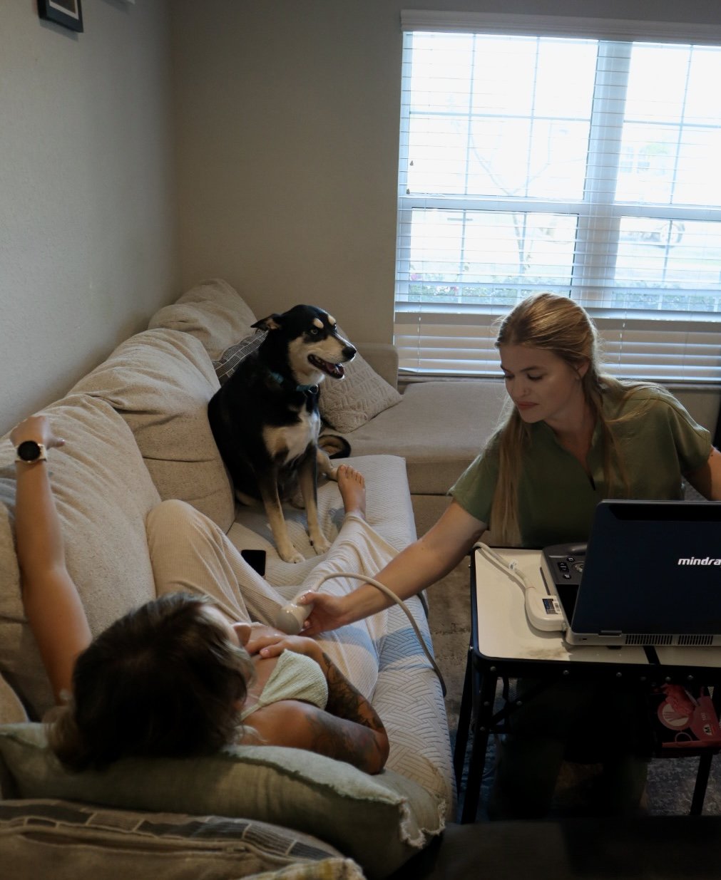 A woman is taking an ultrasound of a woman lying on a couch with her arm raised. A dog is sitting on the couch beside them, and there are blinds on the window behind.