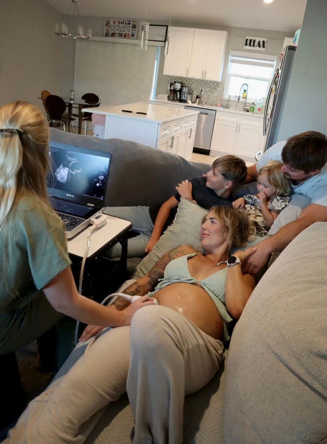 A woman is lying on a couch with an ultrasound probe on her pregnant belly, while a medical professional conducts an ultrasound. Several family members, including children and a man, are gathered around the woman on the couch, watching the ultrasound on a laptop.