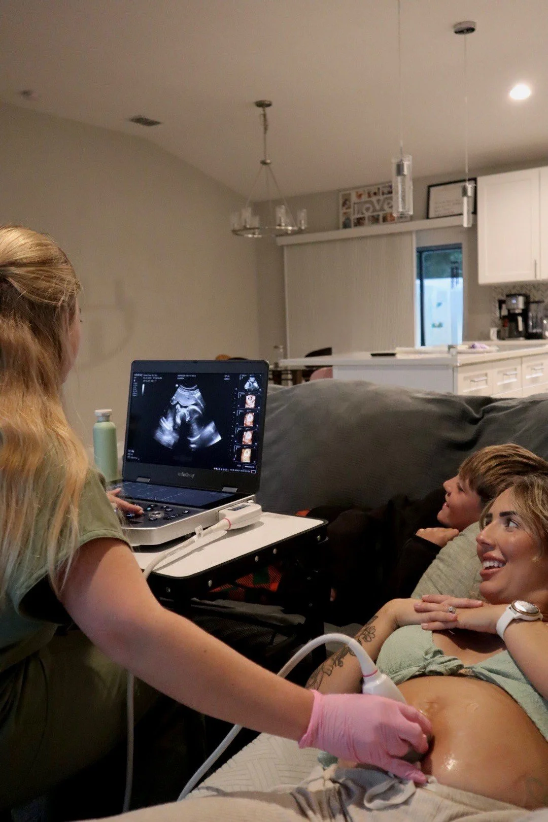 A woman lying on a couch is having an ultrasound scan of her pregnant belly while a healthcare professional examines her with an ultrasound device. A boy sits nearby, watching the screen.