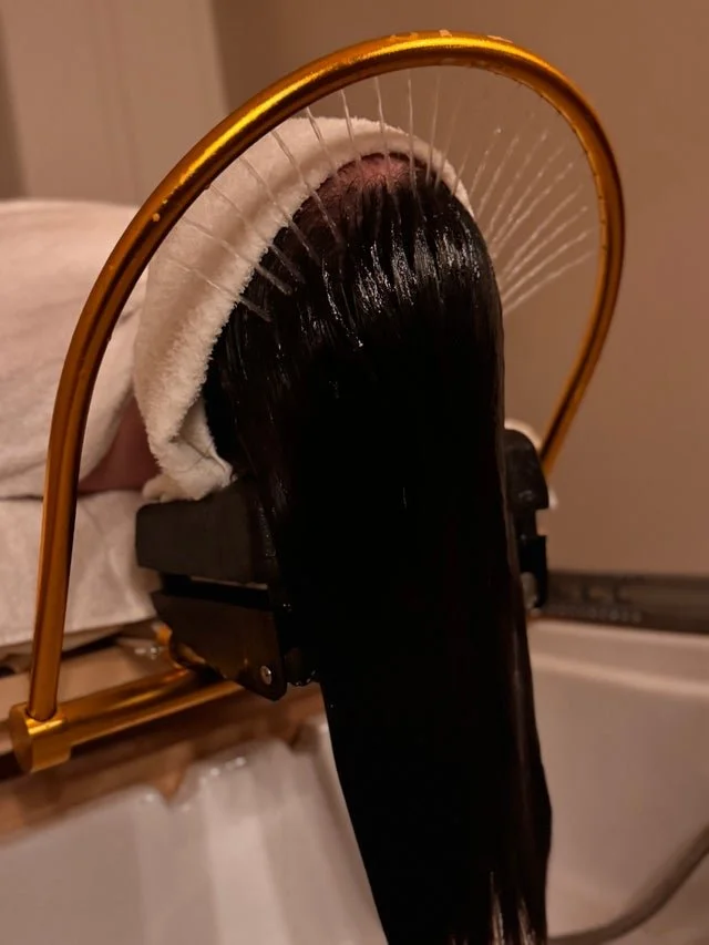 A person lying face down with long, dark hair hanging over a massage or treatment device that has multiple thin strings or needles, with a gold-colored arch above it.