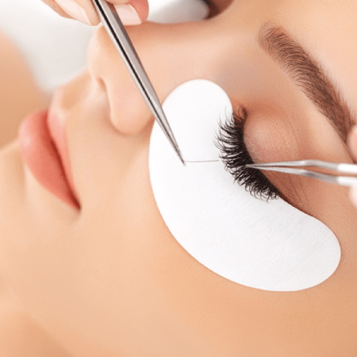 Close-up of a woman receiving eyelash extension procedure with tweezers and adhesive, her eye closed and protected by a protective pad.
