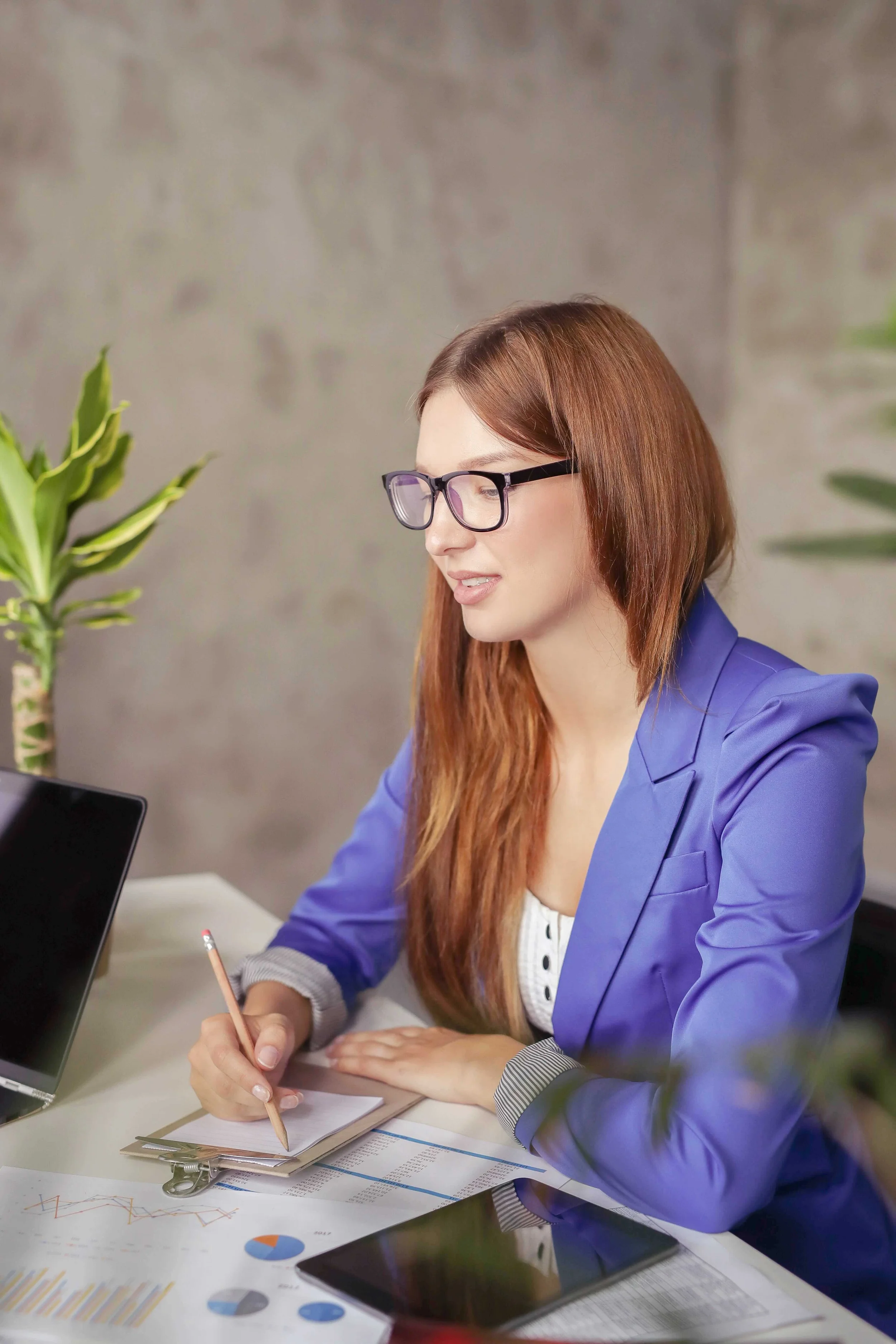 A woman with red hair and glasses working at a desk with documents, a tablet, and a potted plant.