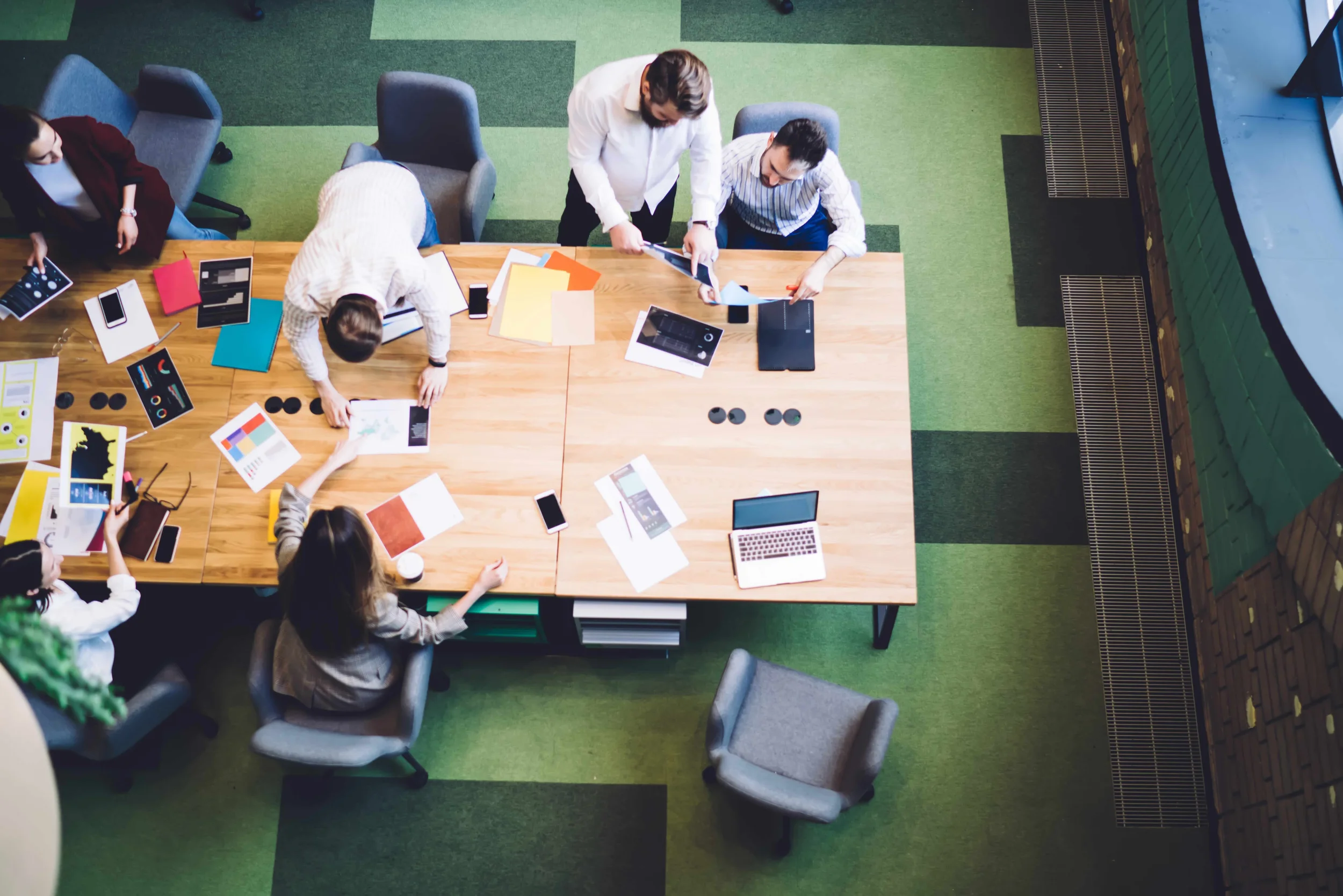 Overhead view of six people in a meeting room working around a large wooden table with laptops, tablets, smartphones, documents, and papers on it. The room has green carpeted flooring and large windows.