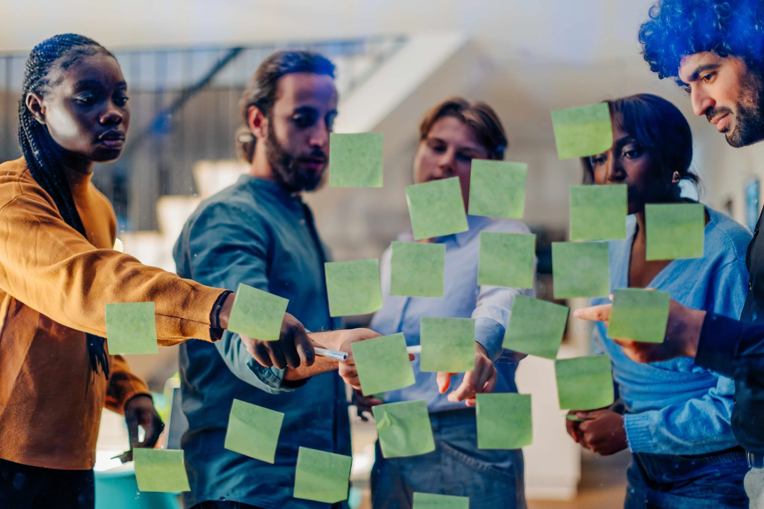 Group of five diverse people in a team brainstorming session, working together with green sticky notes on a transparent glass wall.