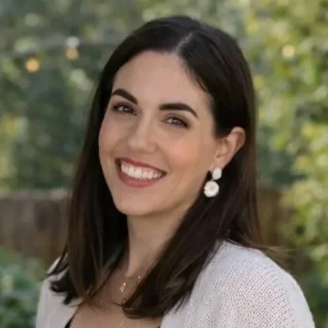 A woman with long dark hair, smiling, outdoors with blurred greenery in the background.