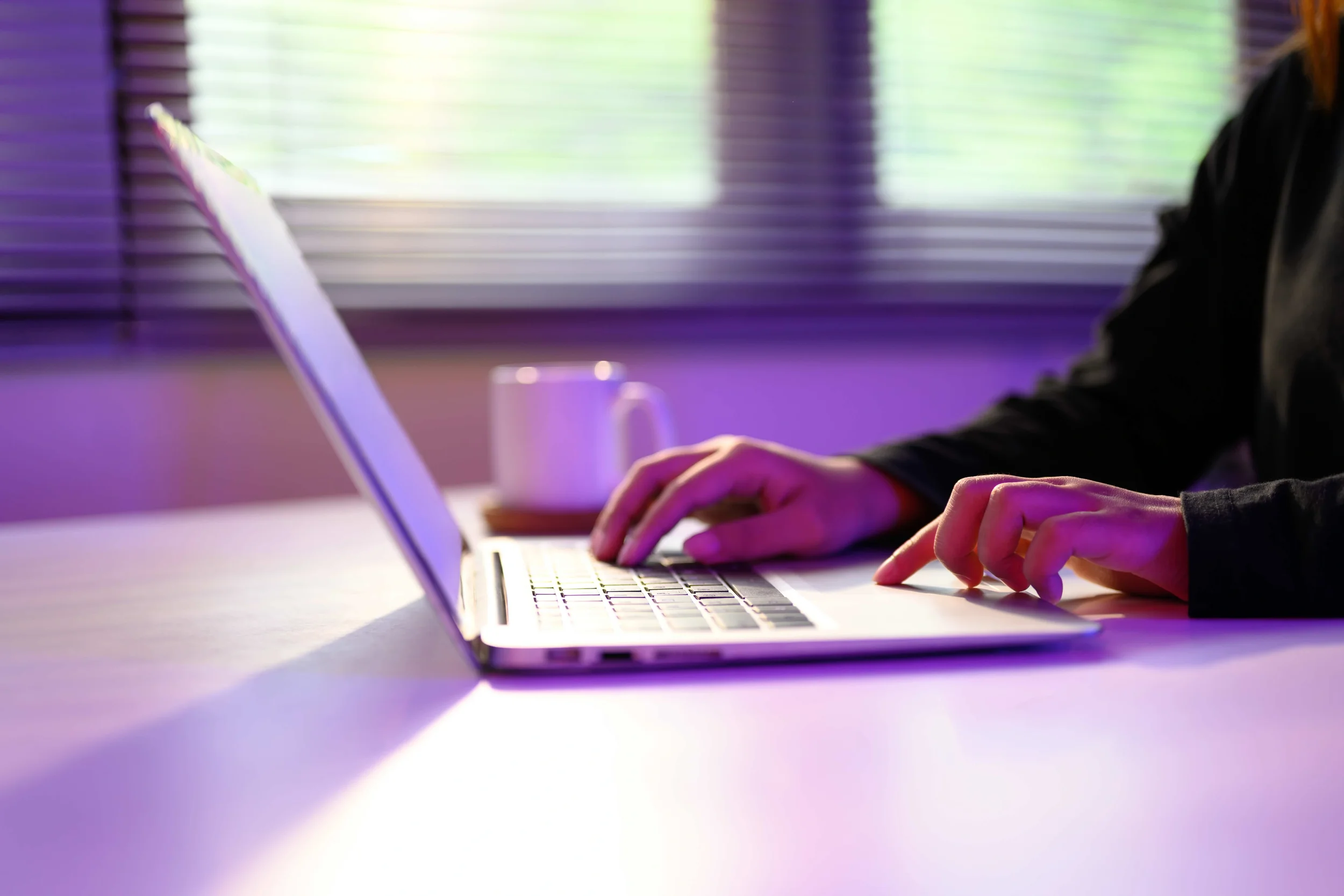 Person typing on a laptop keyboard with a coffee mug in the background, illuminated by purple and green light from a nearby window.