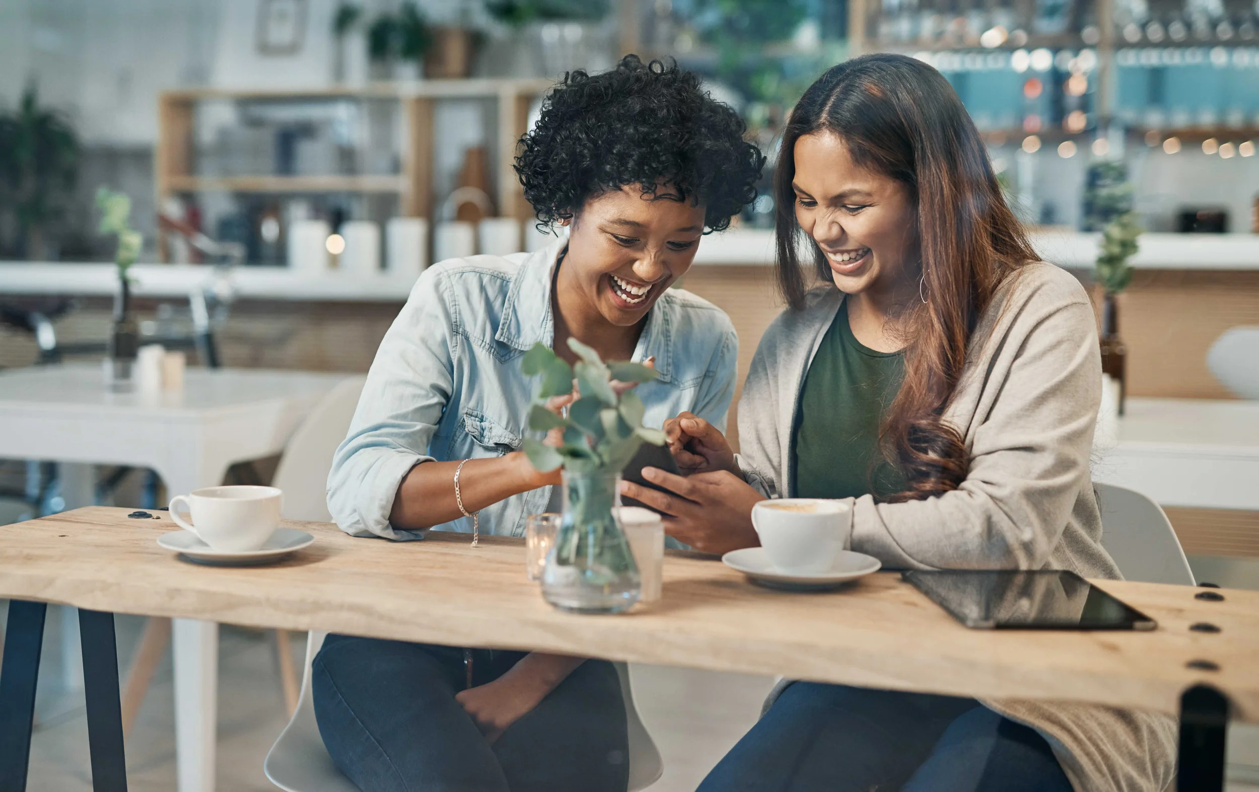 Two women sitting at a wooden table in a cafe, sharing a moment looking at a phone together, smiling and laughing.