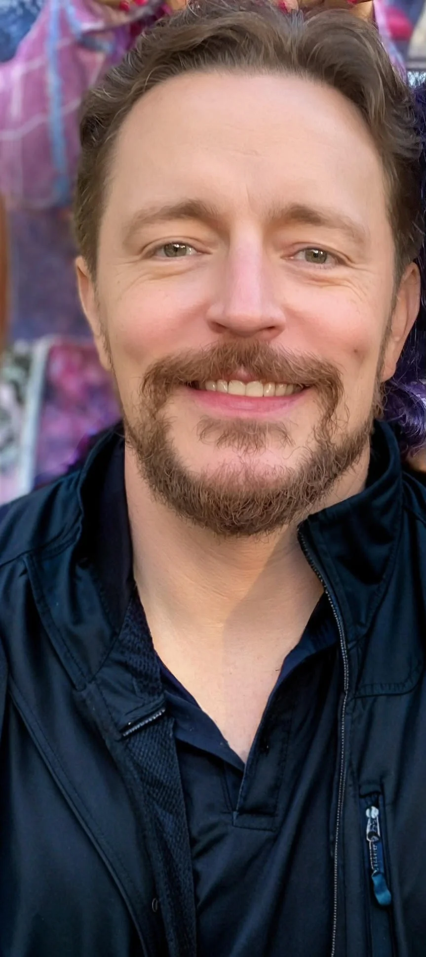Close-up of a man smiling, with light skin, a brown beard, and light-colored eyes, wearing a black jacket.