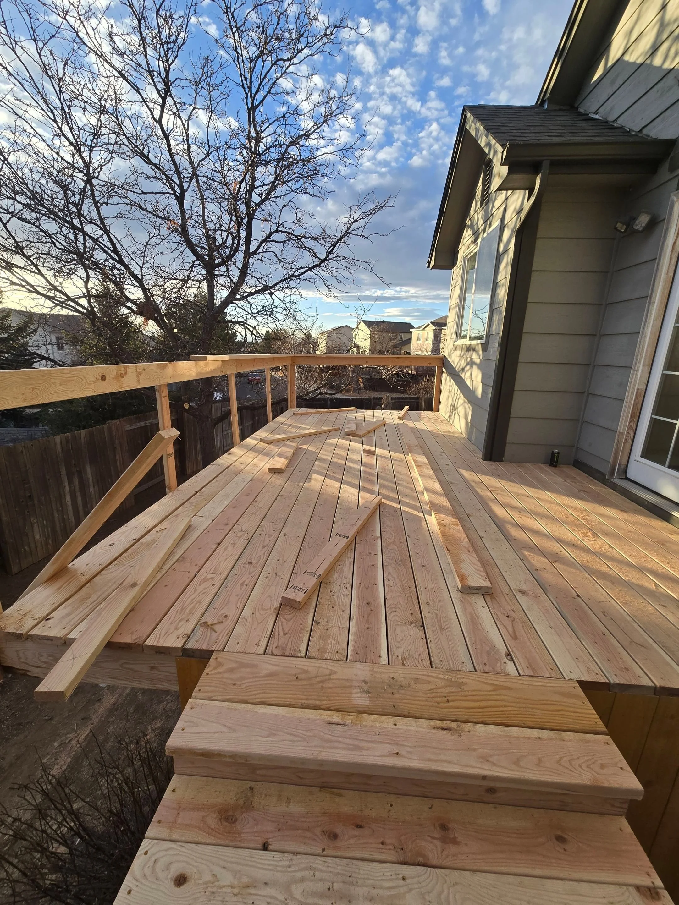 Wooden deck under construction on the back of a house with steps, railing, and tools, surrounded by trees and neighboring houses, under a partly cloudy sky.