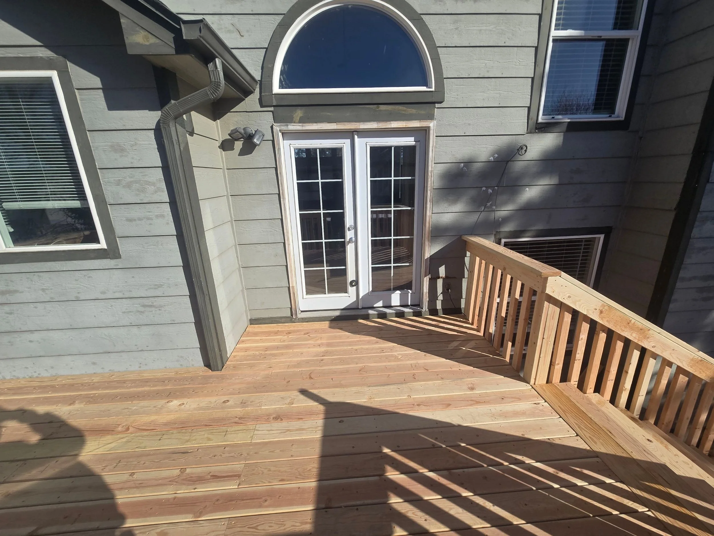 New wooden deck with railing attached to a house exterior, with a glass door and two windows visible, gray siding on the house, and shadows cast by the railing and person taking the photo.