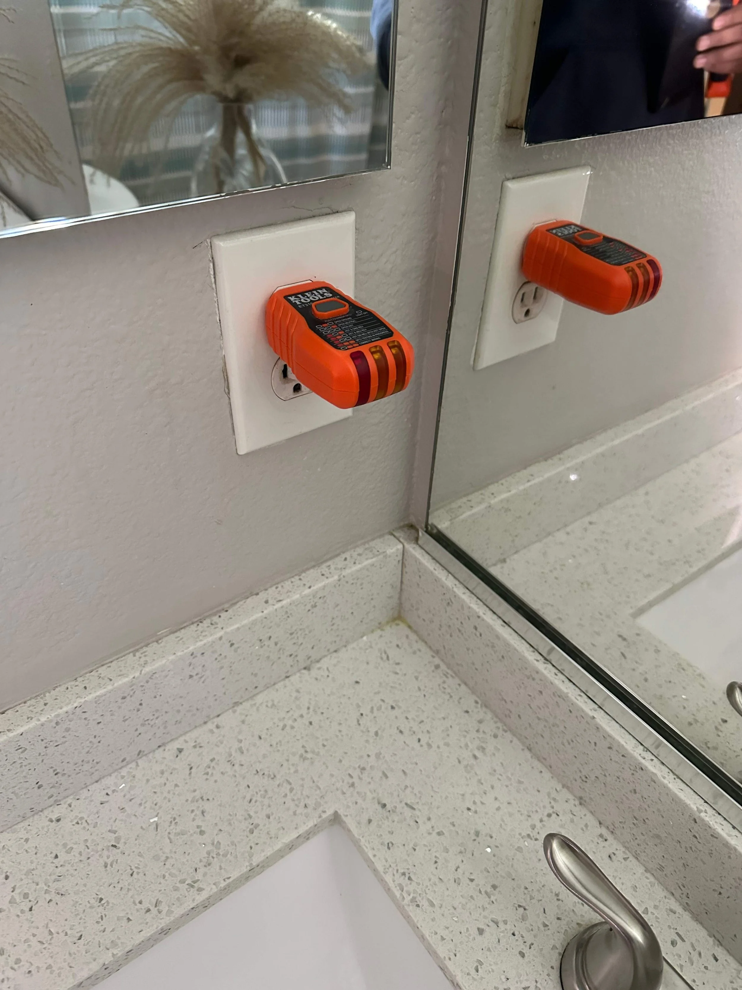 Two orange handheld devices plugged into electrical outlets on a bathroom wall near a mirror and sink.