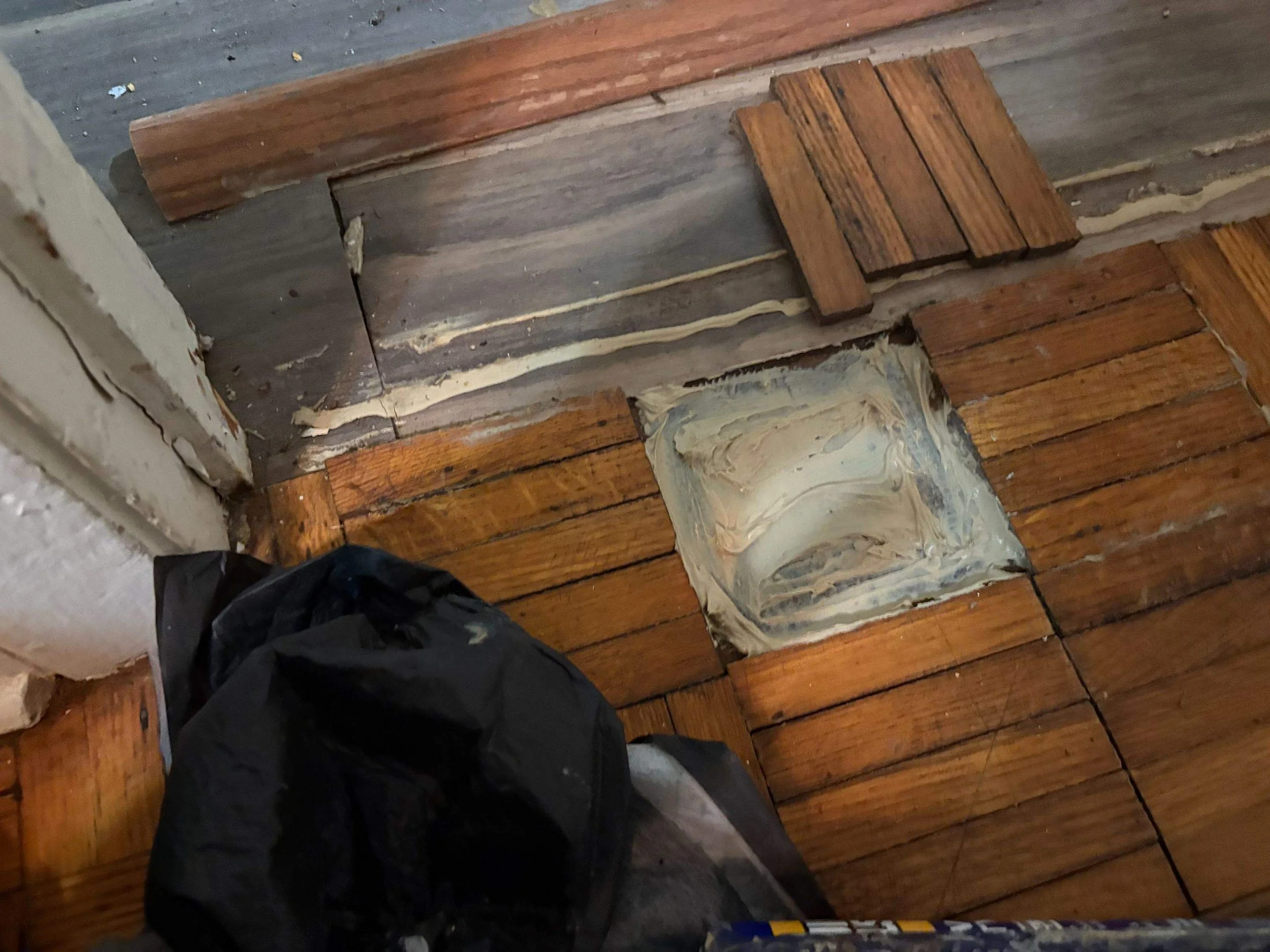 Close-up of a corner of a room showing a wooden floor with some tiles removed, revealing part of the subfloor and a patch of filler or adhesive. A black trash bag and a white wall are also visible.