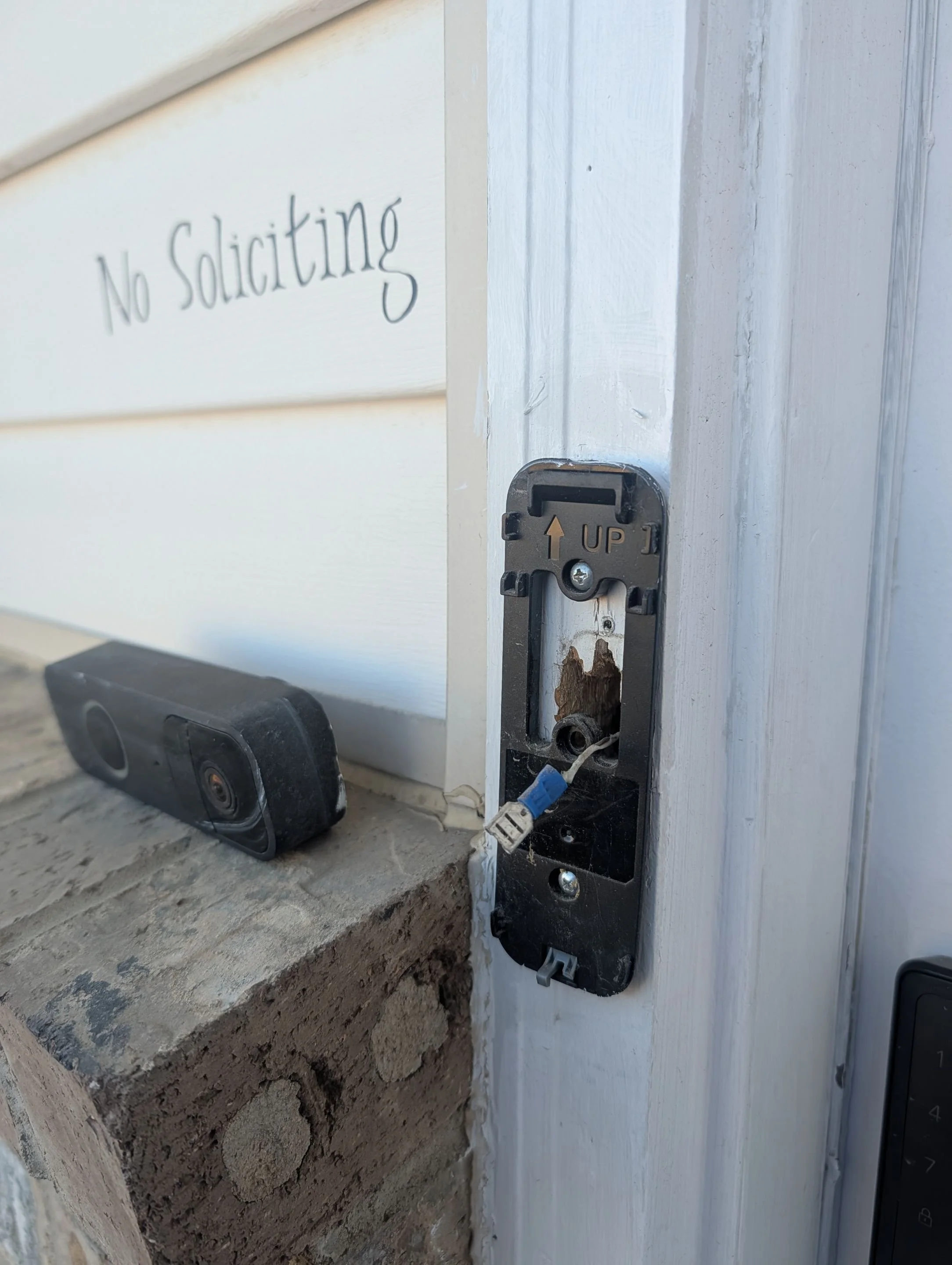 Close-up of a door latch with wires, lacking a handle, next to a white wall with a sign that reads "No Soliciting" and a security camera on a brick surface.