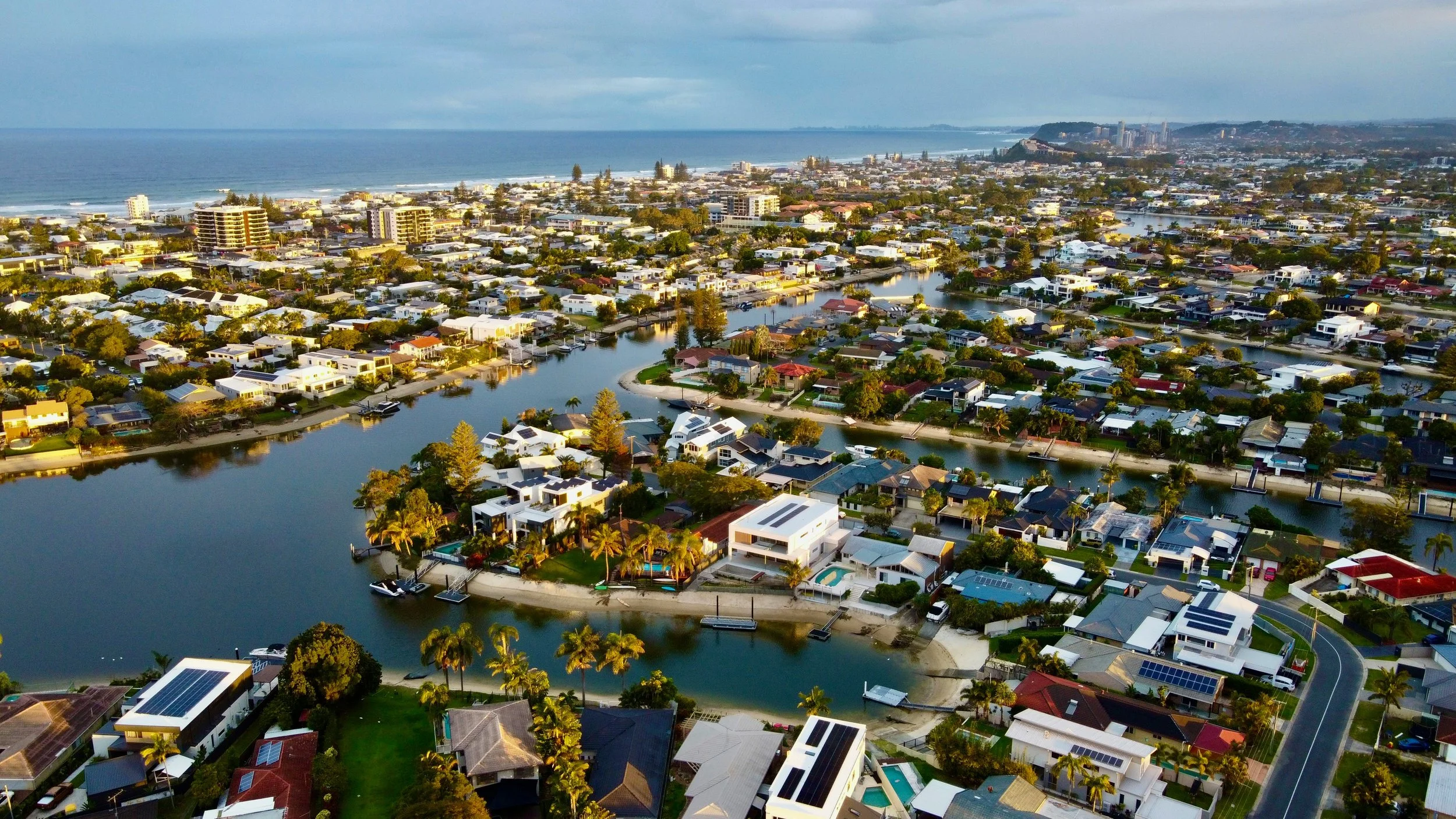 Aerial view of a coastal residential neighborhood with houses along a canal, some with boats, with the ocean and shoreline in the background.