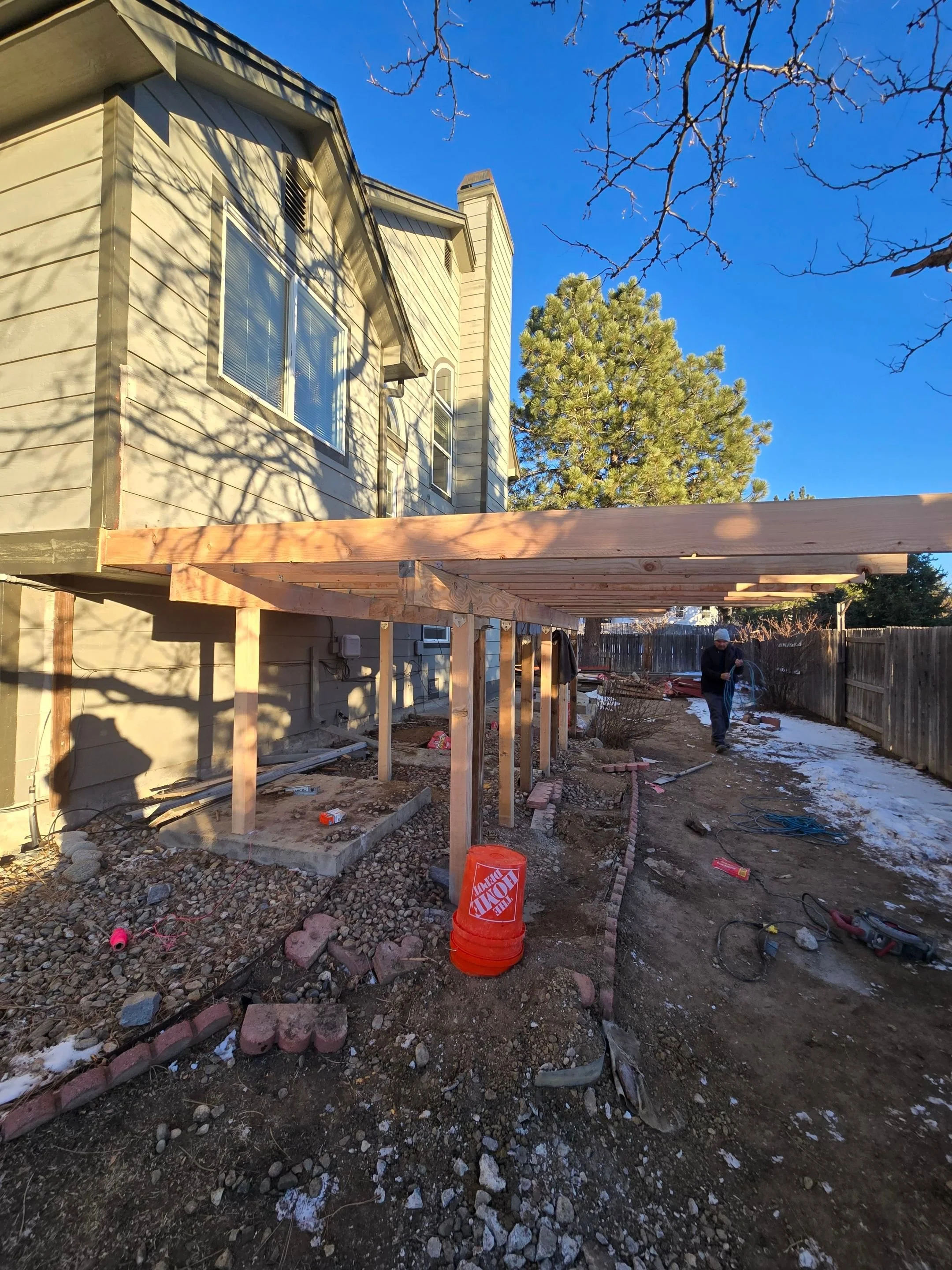 Construction site with a wooden structure being built in backyard, house in background, cloudy sky, and workers present.