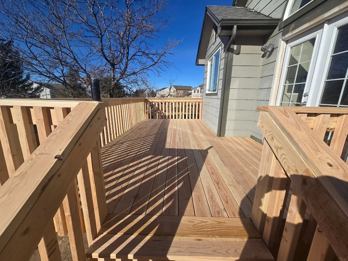 Newly built wooden deck attached to a house with gray siding, clear blue sky, and a leafless tree in the background.