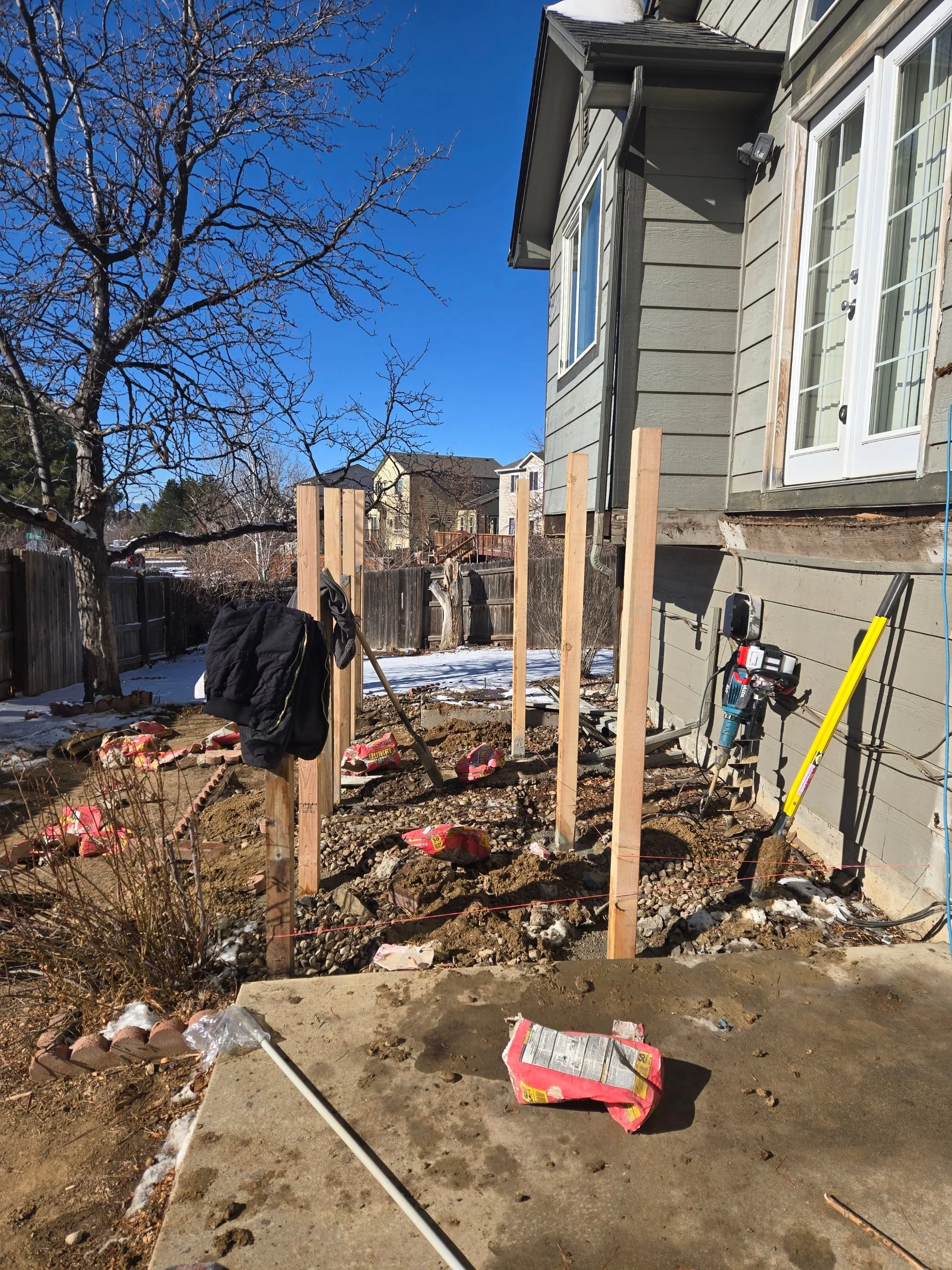 Construction site with wooden posts and tools outside a house, with snow on the ground and a clear blue sky.