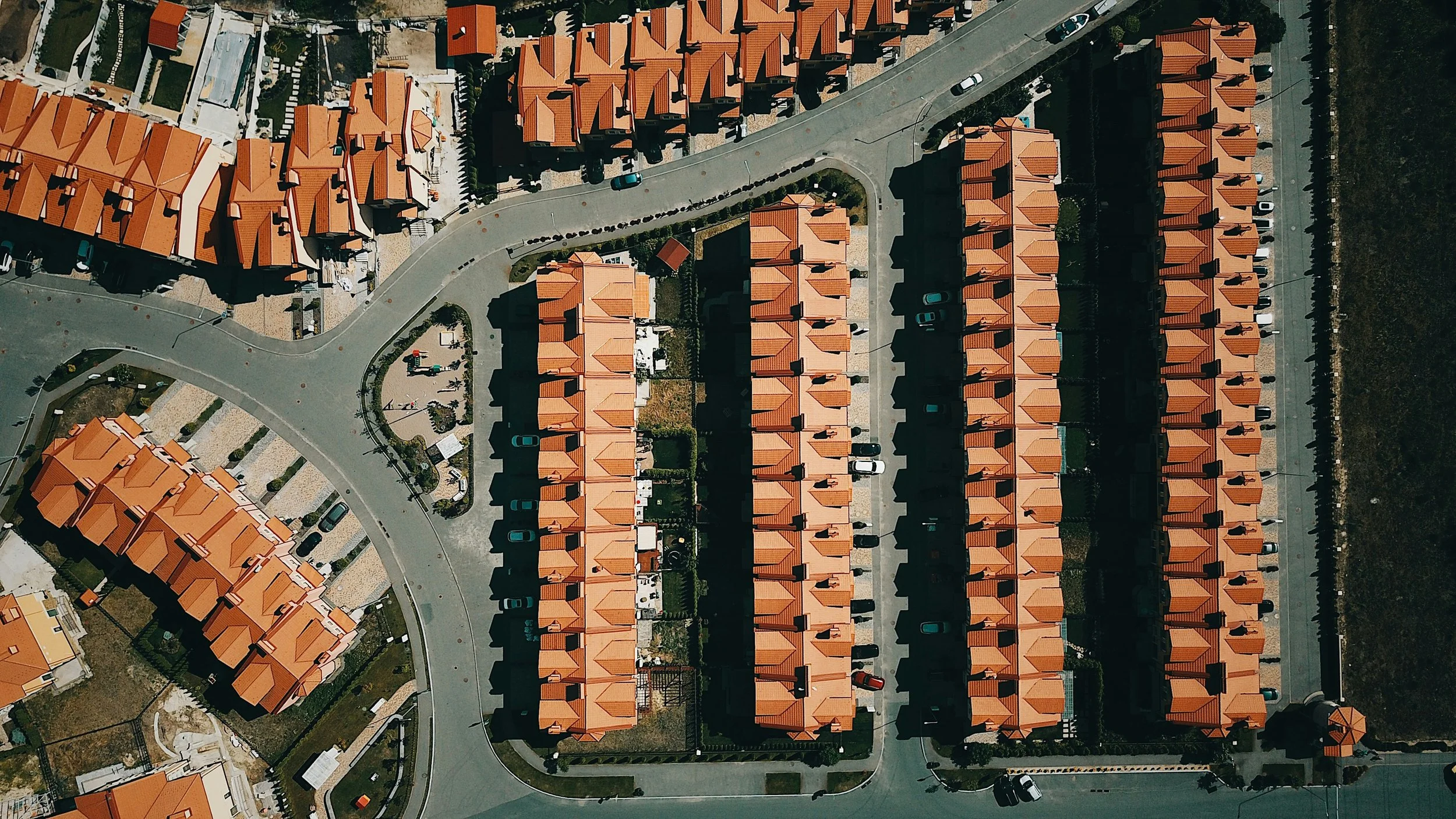 An aerial view of a residential neighborhood showing rows of houses with orange roofs, roads, parked cars, and small yards.