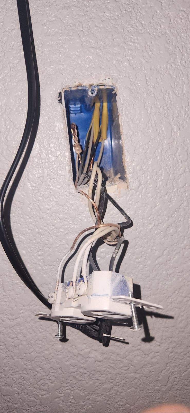 Open electrical outlet box with disconnected wires hanging from a textured ceiling wall.