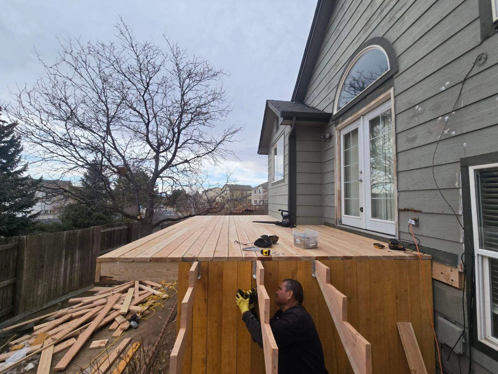 A man working on a wooden deck extension attached to a gray house, with tools and materials on the deck, and a tree without leaves in the background.