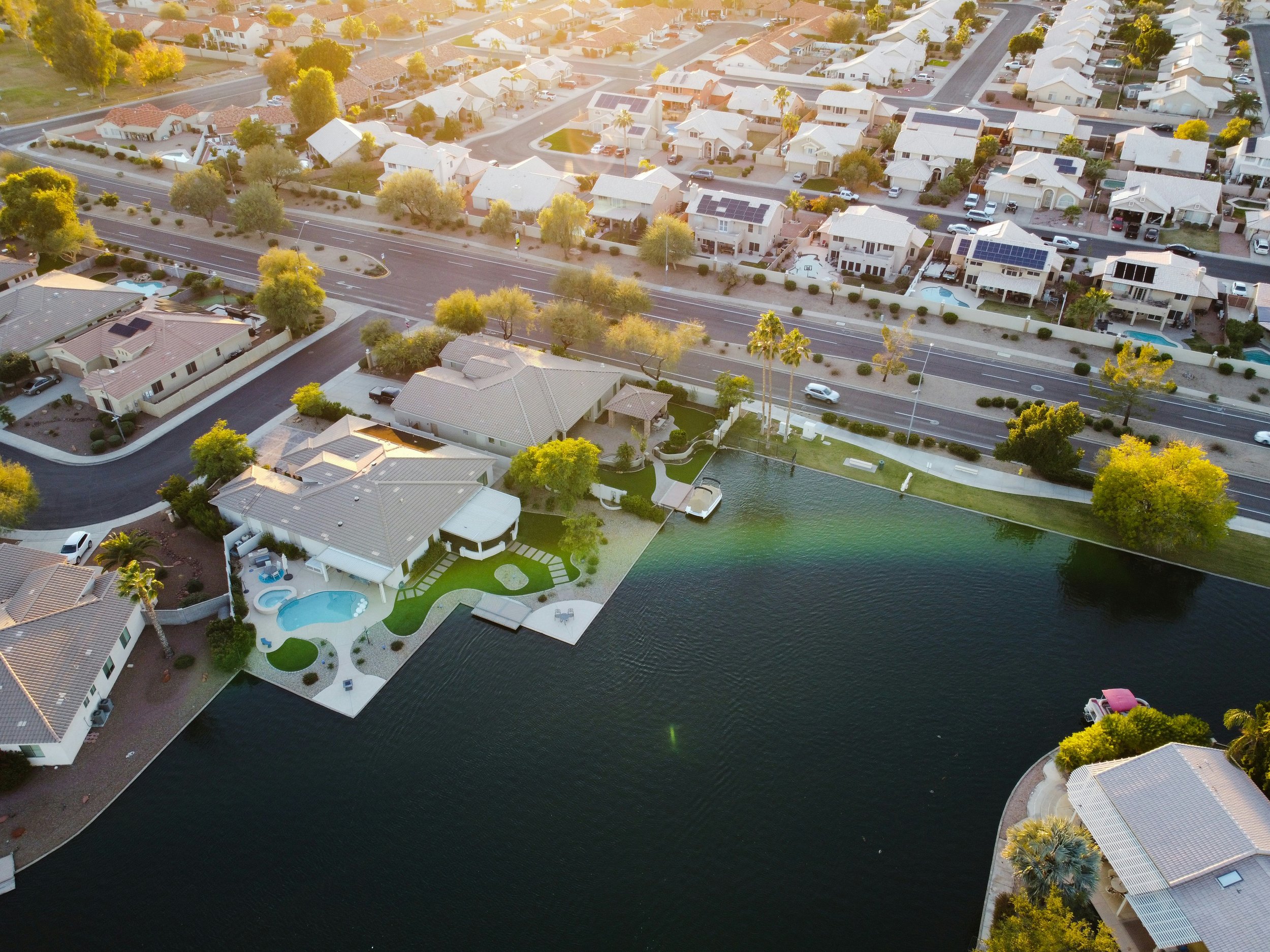 Aerial view of a residential neighborhood featuring houses, trees, and a body of water, with a street running through the area, some houses have solar panels on their roofs, and there are cars parked and driving along the streets.