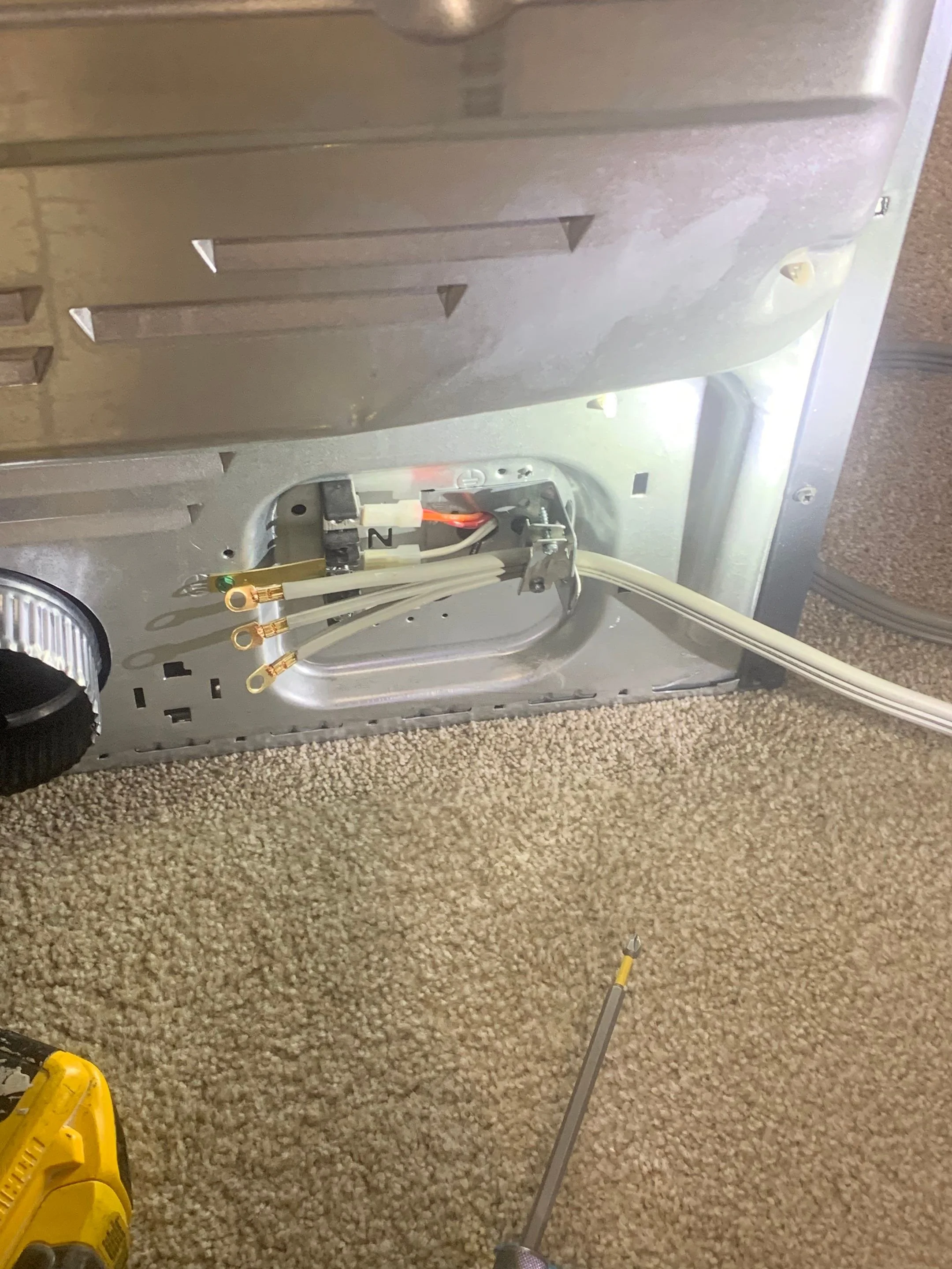 View of the back of a microwave oven with its panel removed, showing internal wiring and electrical connections, placed on a beige carpeted floor next to a yellow screwdriver.